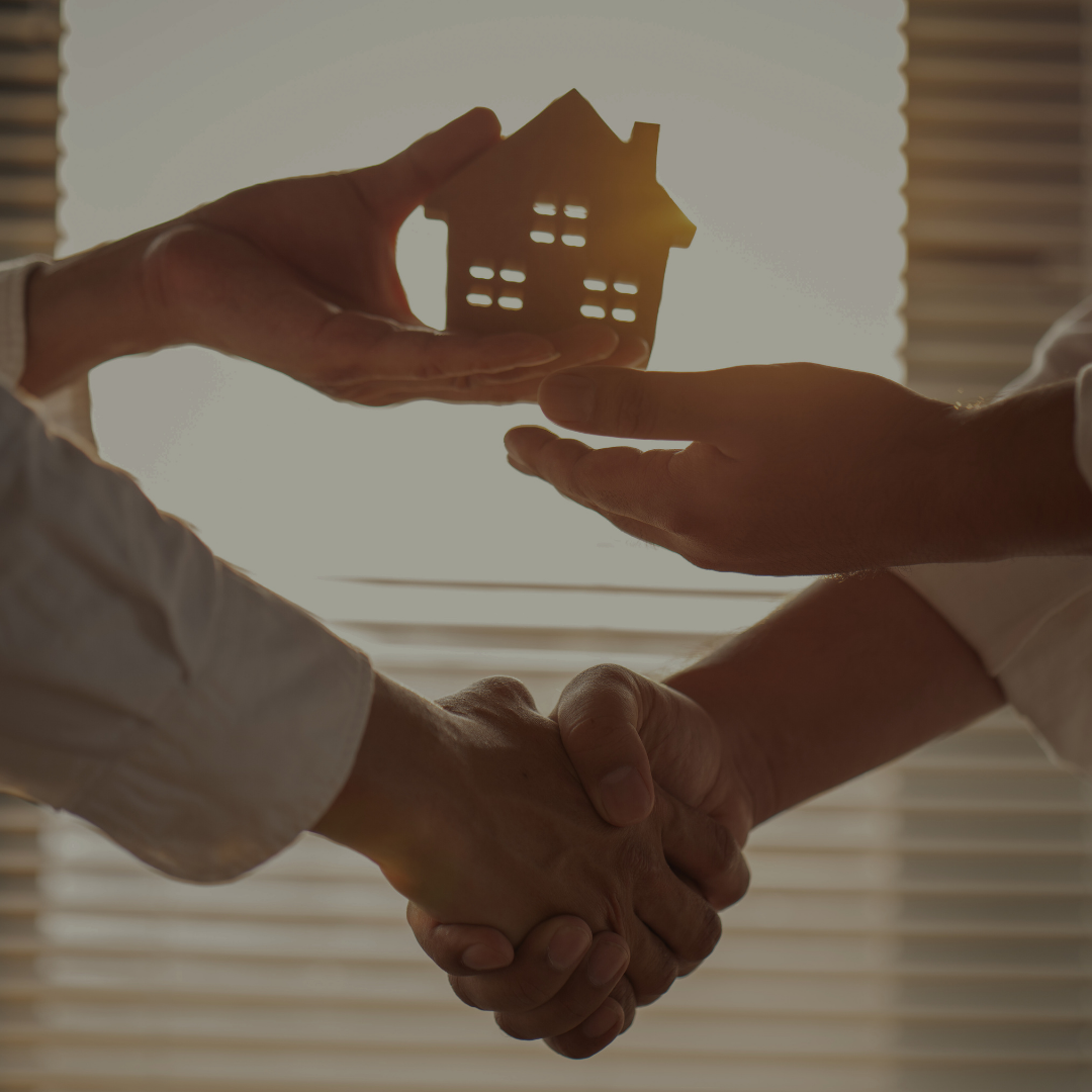 Two people shake hands, with a small house model held between them in front of a backlit window.