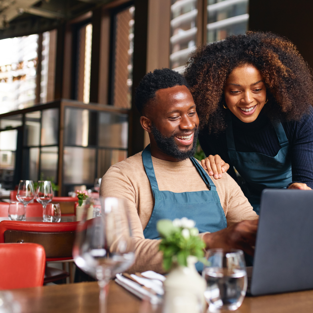 A man and a woman, both wearing aprons, smiling and looking at a laptop in a restaurant with empty tables, wine glasses, and flowers.