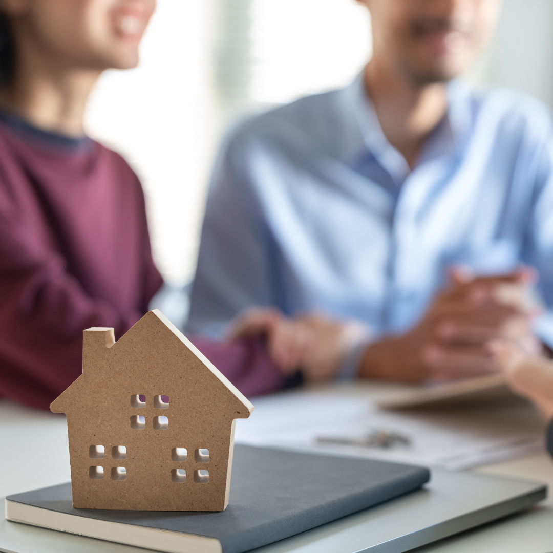 A small cardboard house model on a notebook, with two people in the background discussing, one holding a tablet.