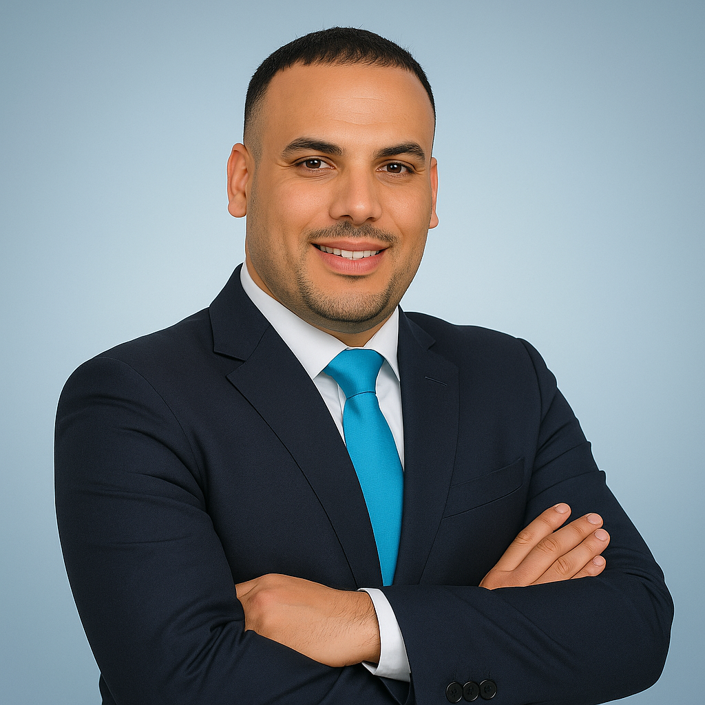 A man in a dark suit and blue tie smiling with arms crossed against a light blue background.