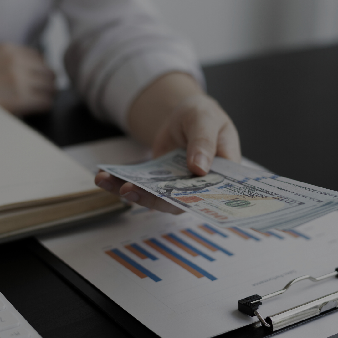 A person's hand handing over a hundred-dollar bill over a desk with financial documents and a bar graph.