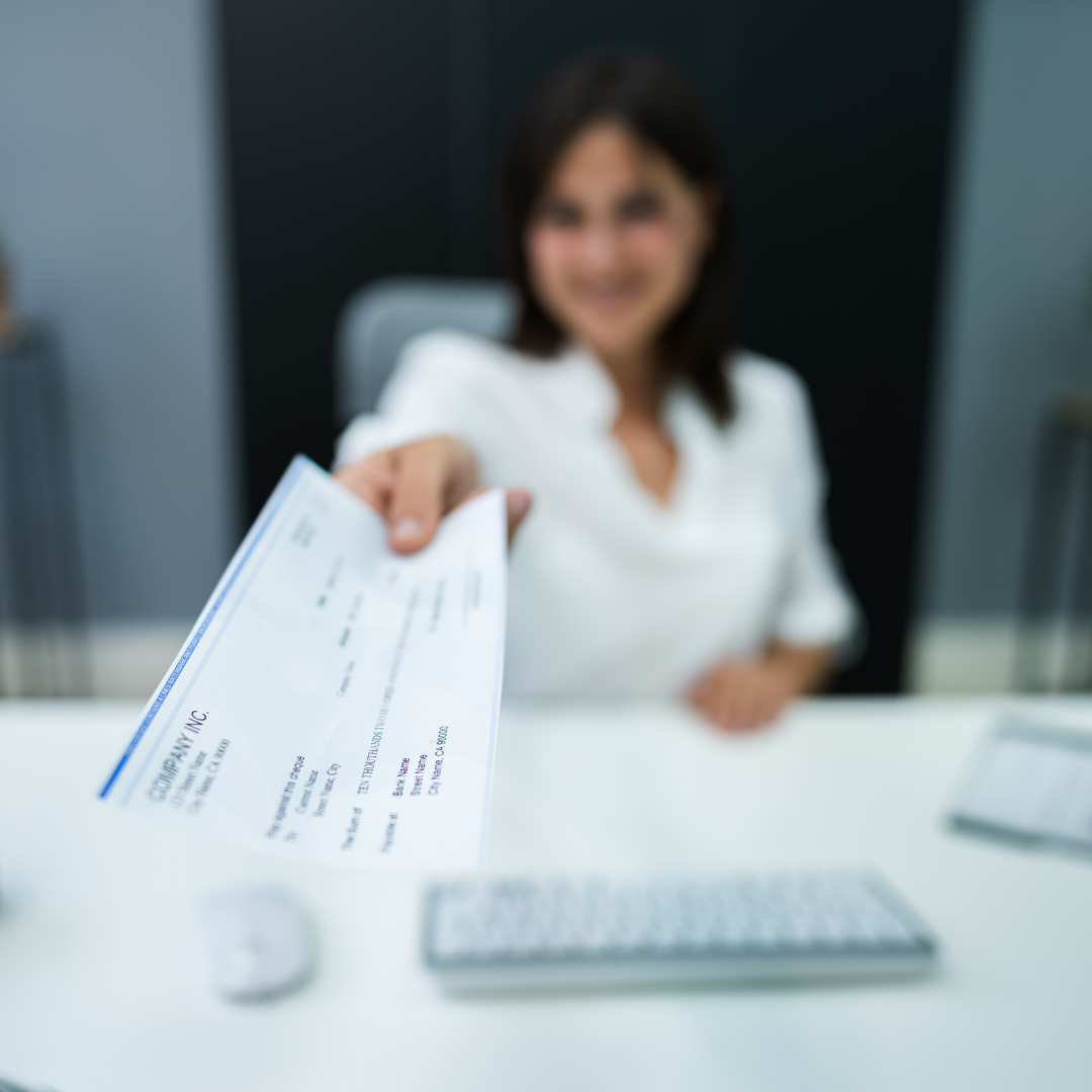 A woman in a white shirt sitting at a desk, reaching out to give a check to the camera.