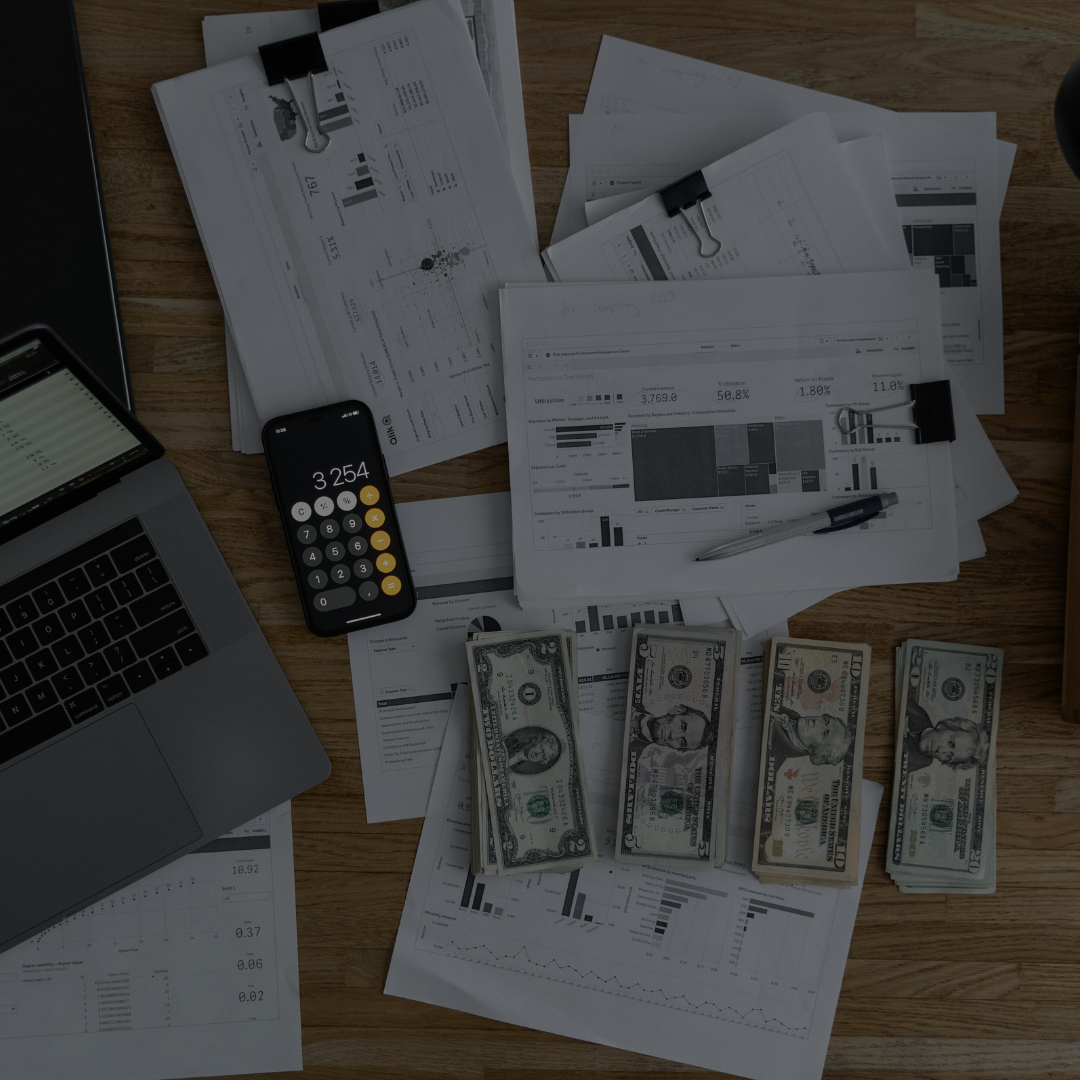 Work desk with papers, graphs, and charts, a laptop, a calculator displaying 3,254, stacks of U.S. dollar bills, a pen, and binder clips on a wooden surface.