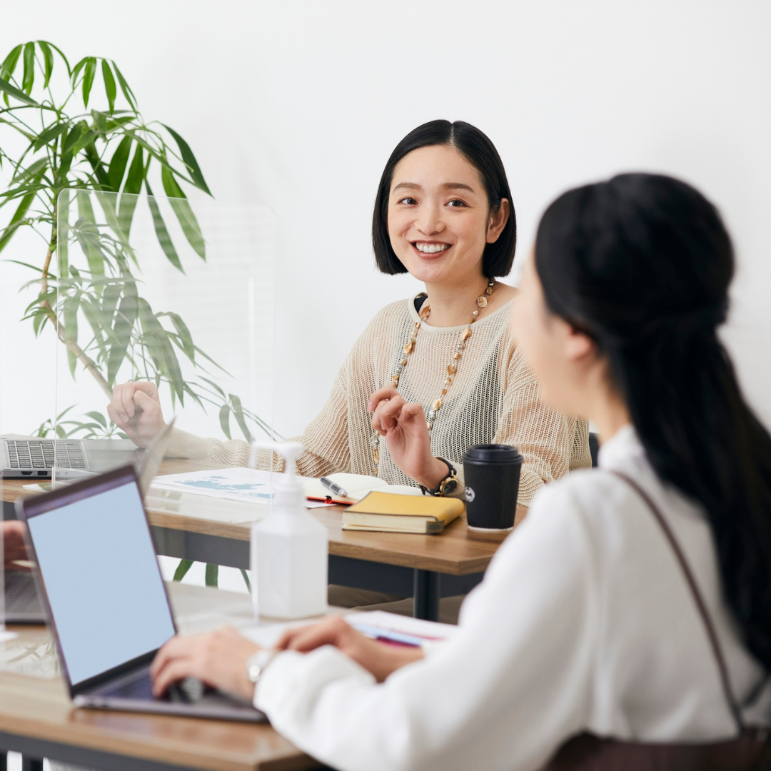 Two women having a conversation during a meeting in an office, with laptops, notebooks, a coffee cup, hand sanitizer, and a large green plant in the background.
