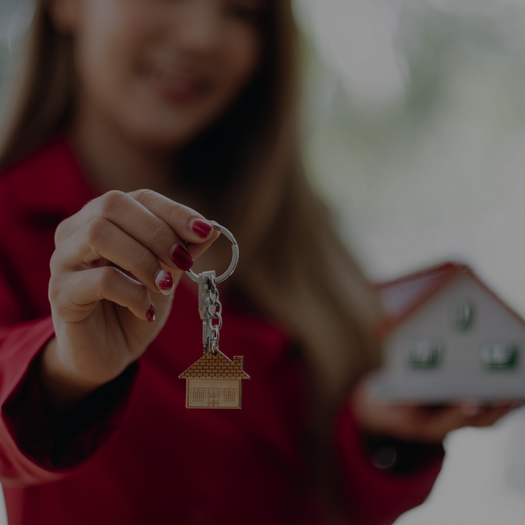Person holding a key with a house-shaped keychain, with a model house in the background.