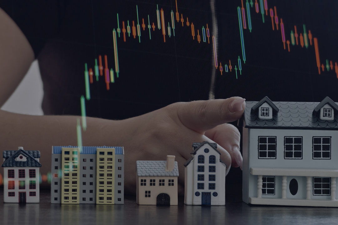 A person holding a tablet with a stock chart in the background, with small house models and buildings on a table in front.