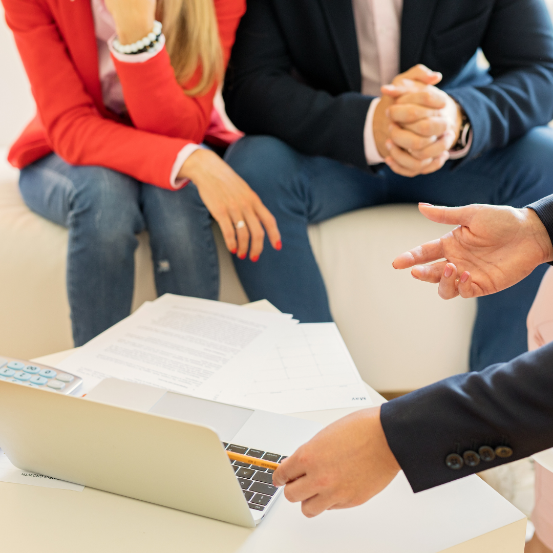 Three people in a business meeting, with one person pointing at a laptop, and papers on the table.