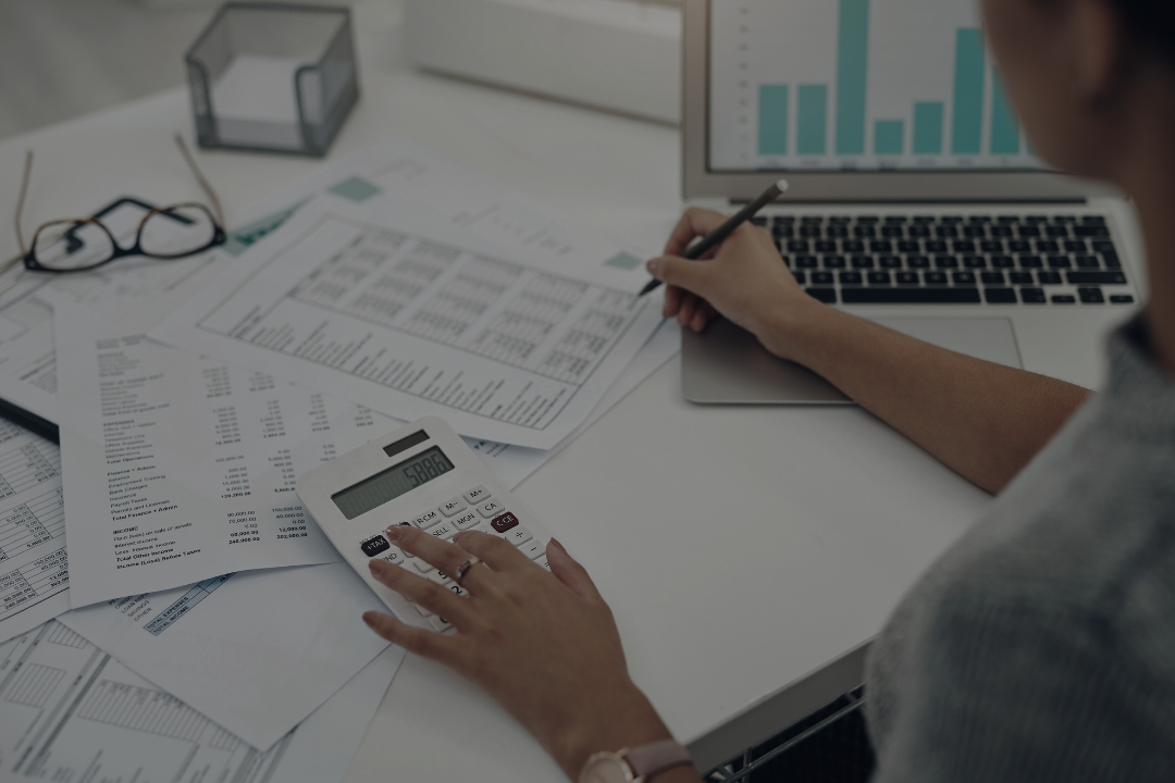 Person working with financial documents, using a calculator, writing on paper, and a laptop with bar graph on the screen.