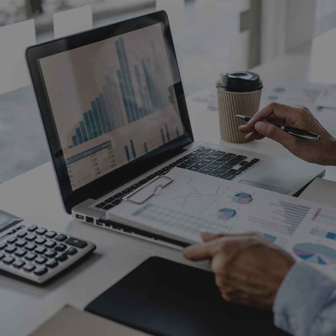 Person analyzing financial charts and reports on a laptop and paper in an office setting.