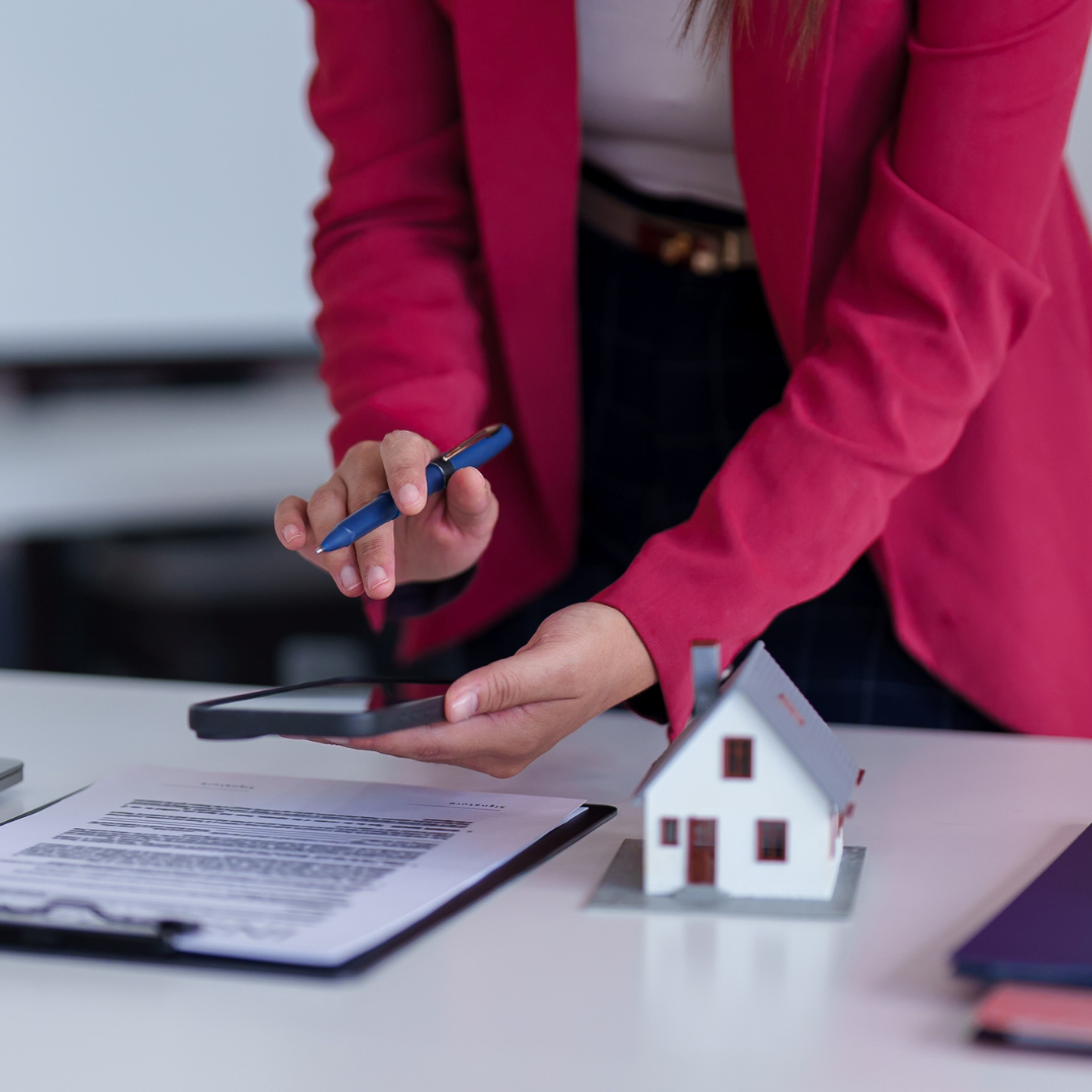 A person in a pink blazer and dark trousers holding a pen and a smartphone, with a small model house, papers, and a purple notebook on a white desk.