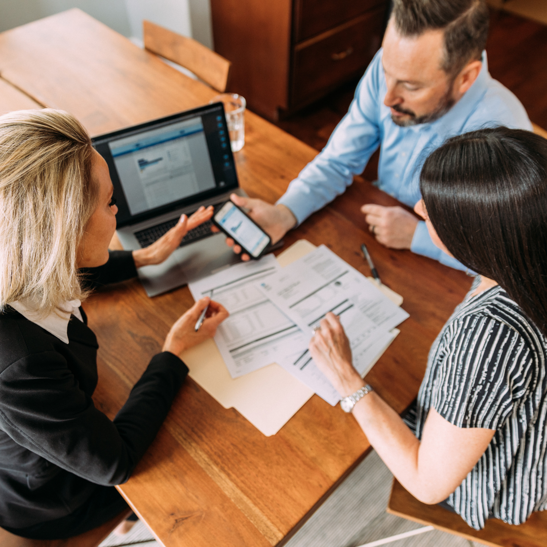 Three professionals in a business meeting around a wooden table with documents, a laptop, and smartphones.