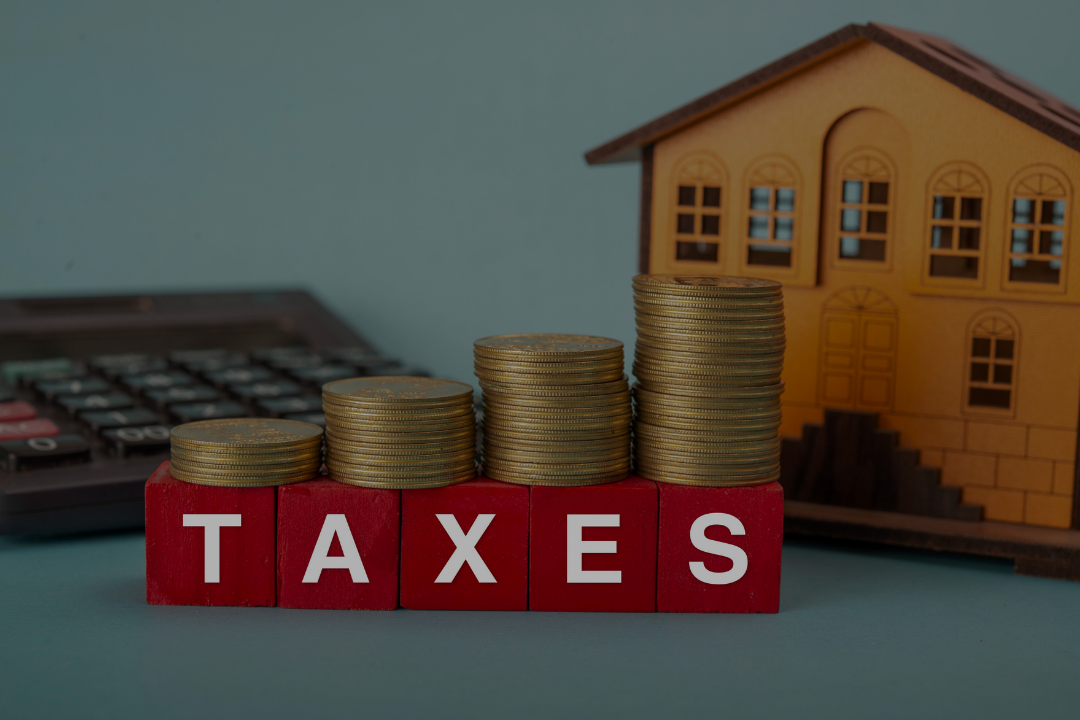 Stacks of gold coins increasing in size in front of red blocks spelling 'TAXES', with a calculator and a small wooden house in the background.