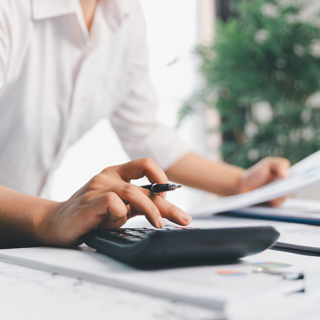 Person in a white shirt calculating with a calculator, holding a pen, and reviewing documents on a desk with papers and charts.