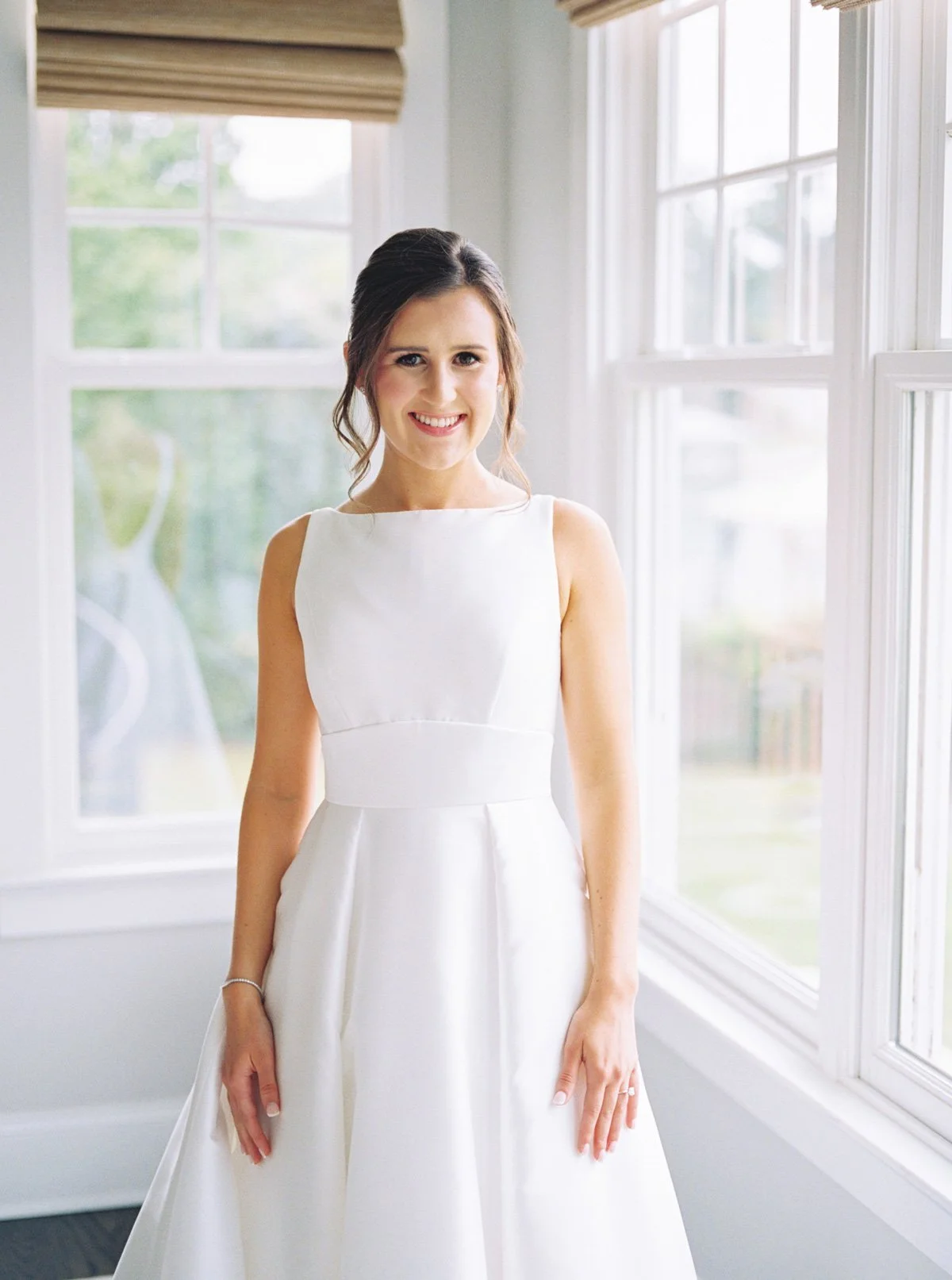 Bride in sleeveless gown posing by window with fresh and polished bridal makeup look
