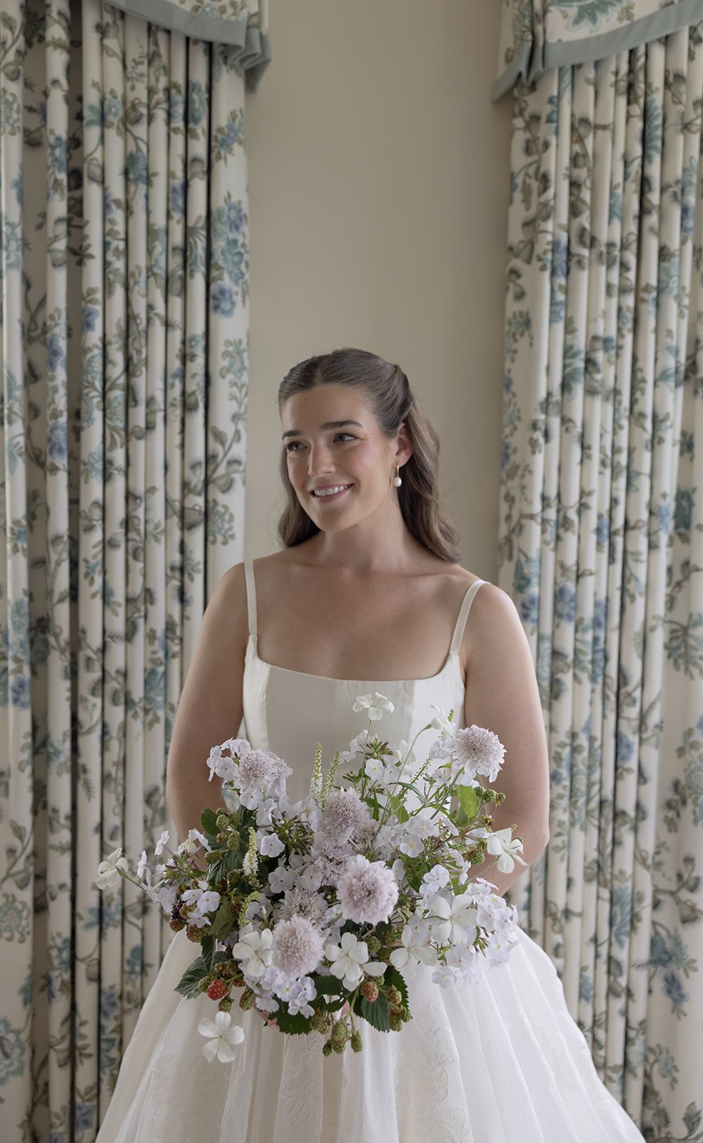 A bride in a white dress holding wild flowers has her hair and makeup done. Her hair is a beautiful half up half down bridal hairstyle. Her bridal makeup is a natural bridal makeup look that enhances her features.
