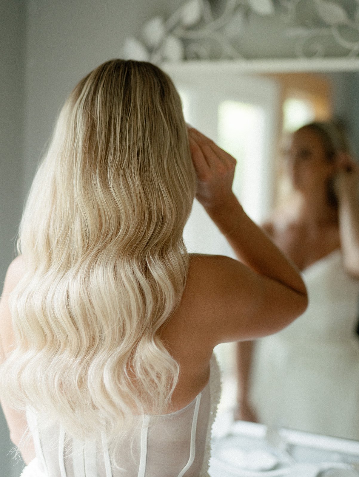 A woman with blonde hair styled in glam waves and wearing a white wedding dress is looking at herself in the mirror admiring her hair. Bridal hair and makeup by professional makeup artist, Carly Amico of Carly Marie Artistry, a makeup artist in Charl