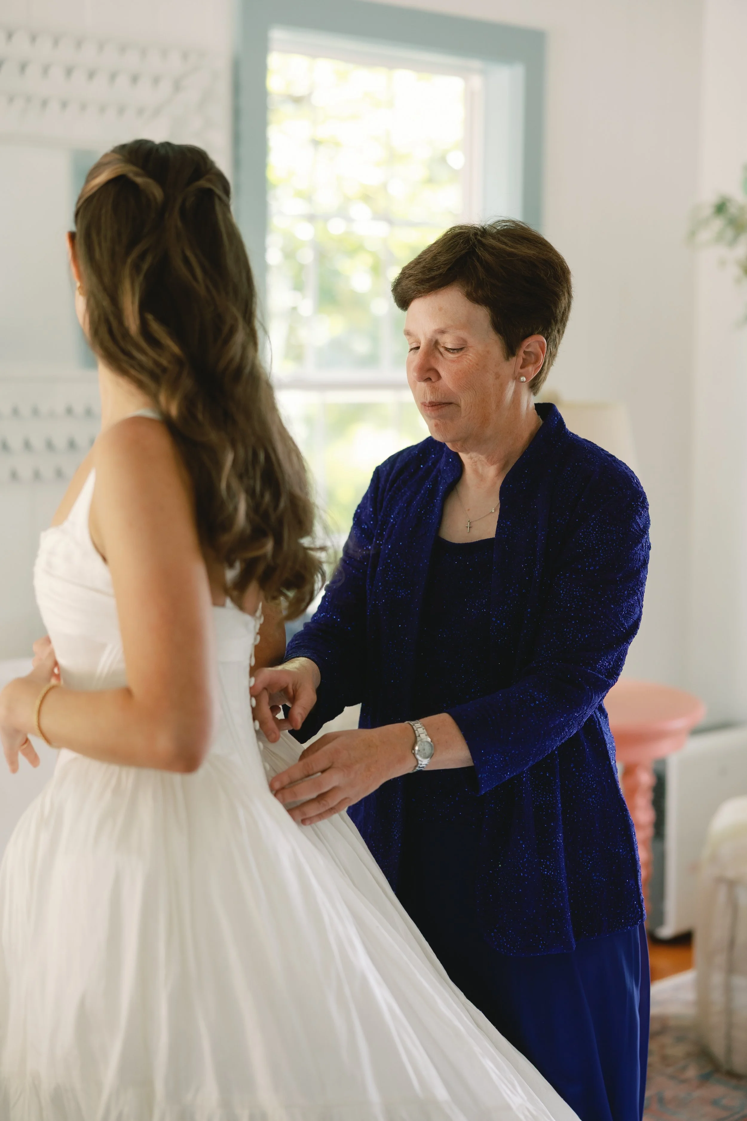 A woman with brown wavy hair is getting her wedding dress buttoned by her mom. Her hair is styled in a bridal half up hairstyle. Bridal hair and makeup by professional makeup artist, Carly Amico of Carly Marie Artistry, a makeup artist in Charlotte N