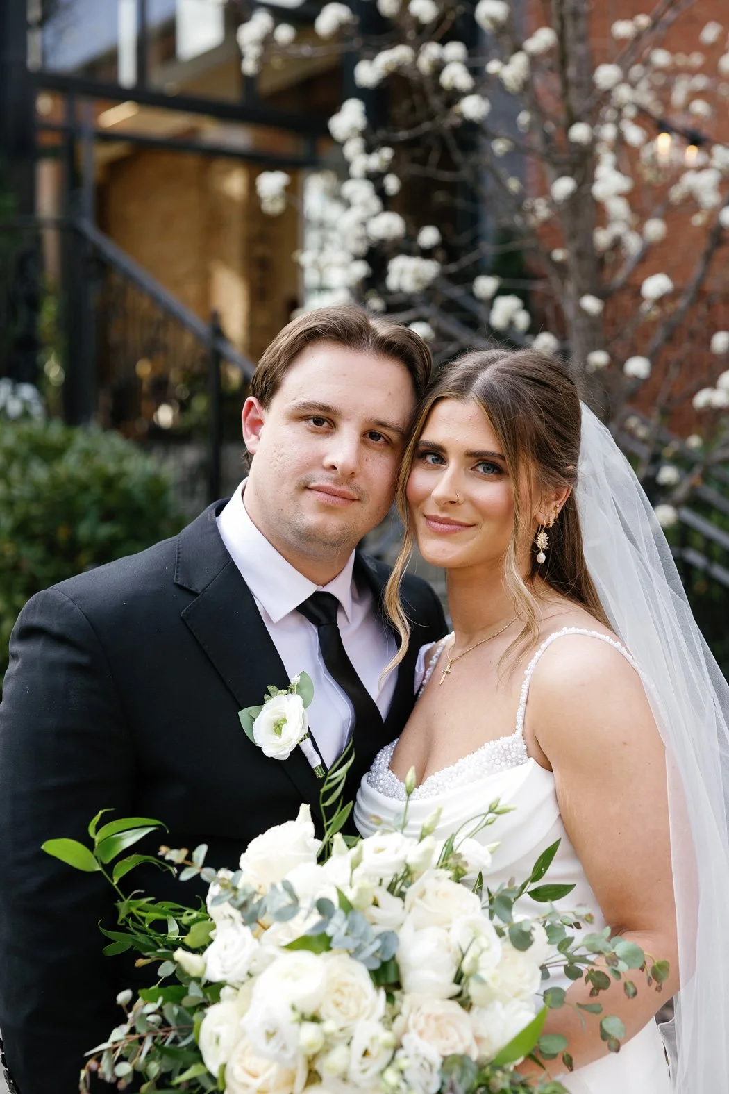 A newlywed couple poses outdoors, the groom in a black suit with a white shirt and black tie, and the bride in a white wedding gown with a veil. The bride holds a large bouquet of white flowers. They stand close together with a blooming tree and building in the background.