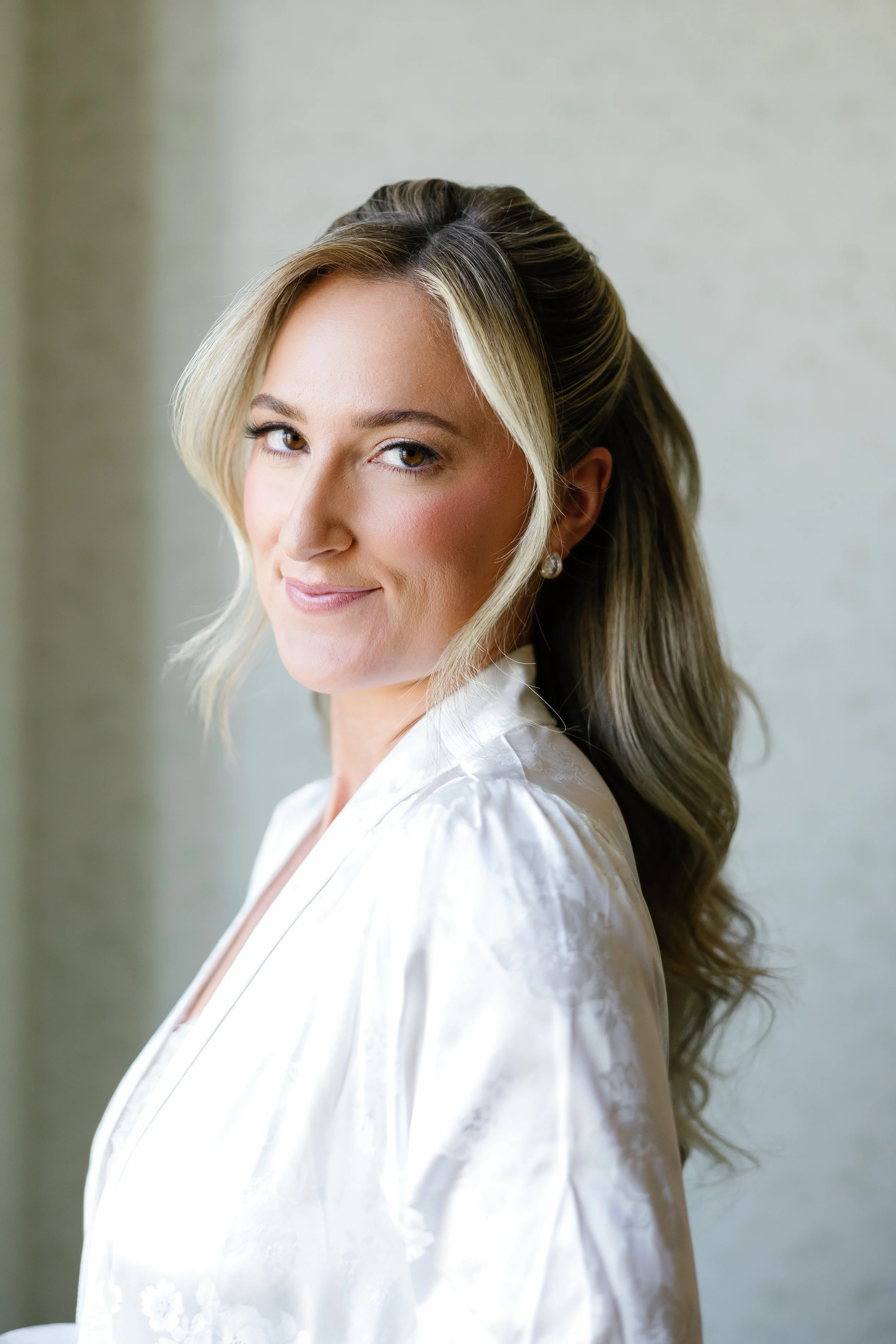 A bride poses in her getting ready robe. Her makeup is a soft glam bridal makeup look that enhances her natural features