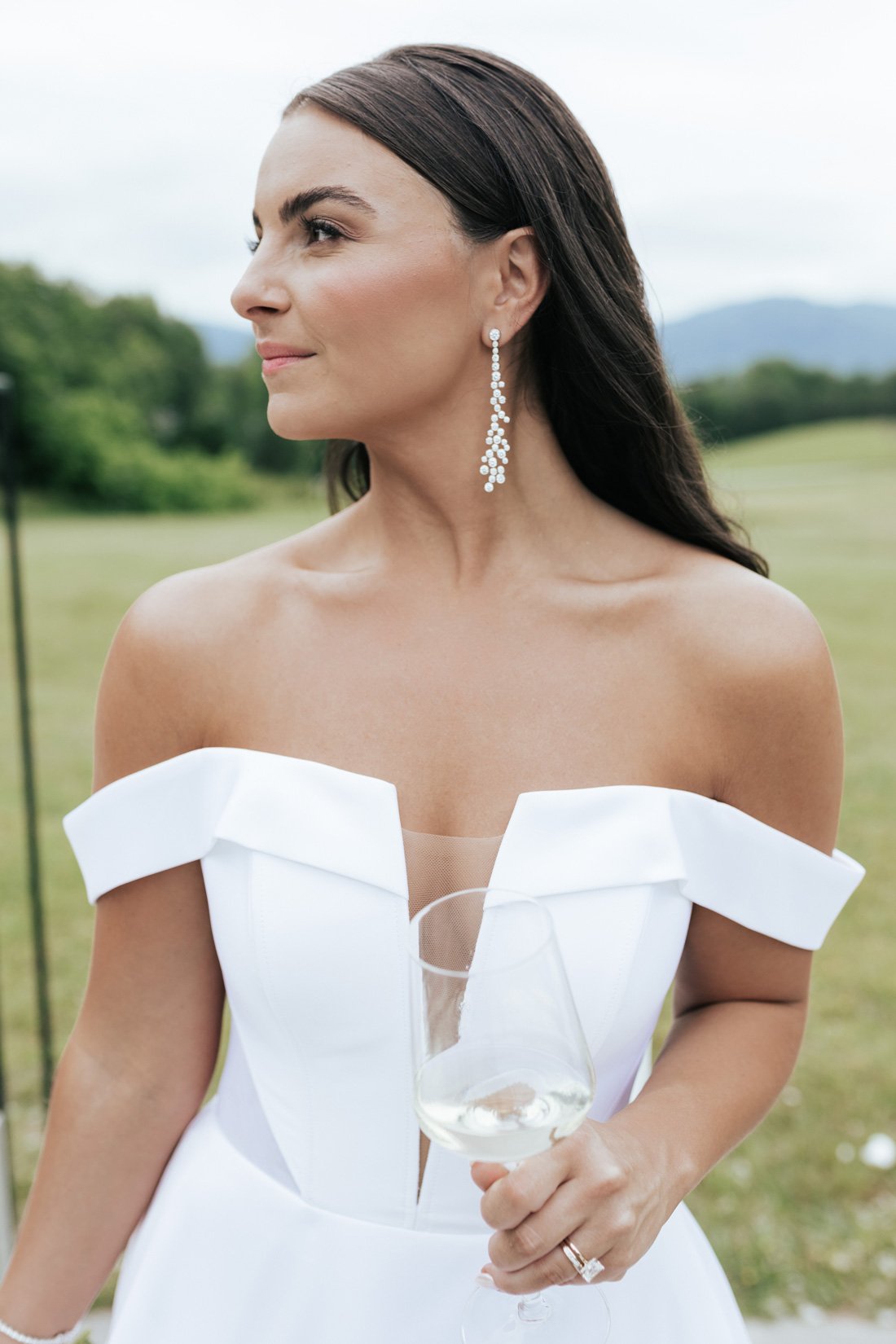 A woman in a white off-shoulder dress, holding a glass of white wine, standing outdoors with a blurred green landscape background.