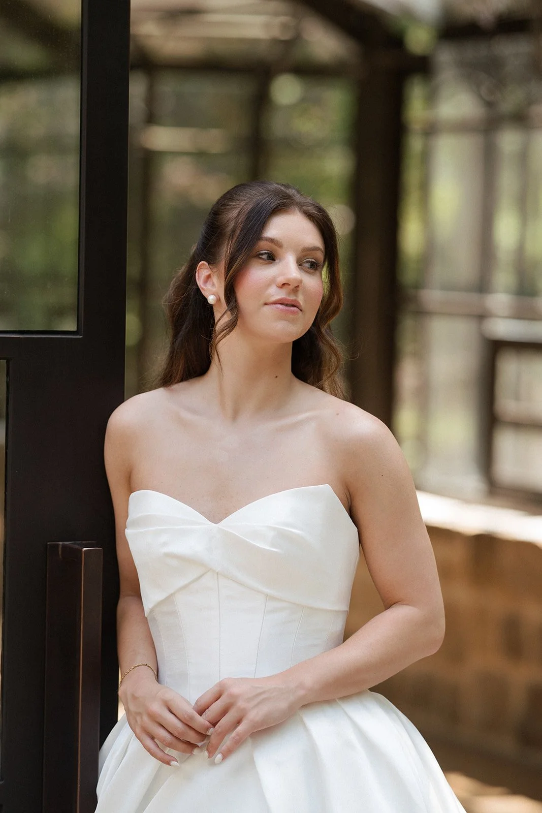 A woman in a white basque waist wedding dress is posing in front of morning glory farms for a bridal photo. Her hair is a half up half down bridal hairstyle with voluminous curls. Her makeup is a soft glam bridal makeup look that looks natural. Brida