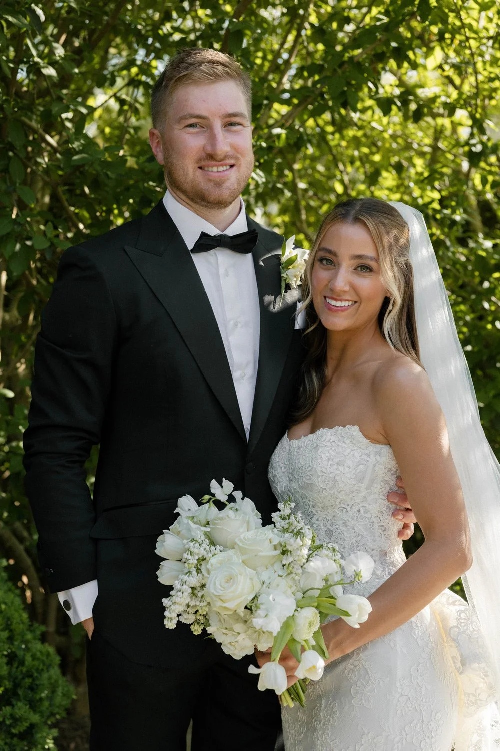 A newlywed couple on their wedding day, standing outdoors in sunlight with green foliage in the background. The groom is in a black tuxedo with a bow tie, and the bride is in a white lace wedding dress holding a bouquet of white flowers, wearing a veil.