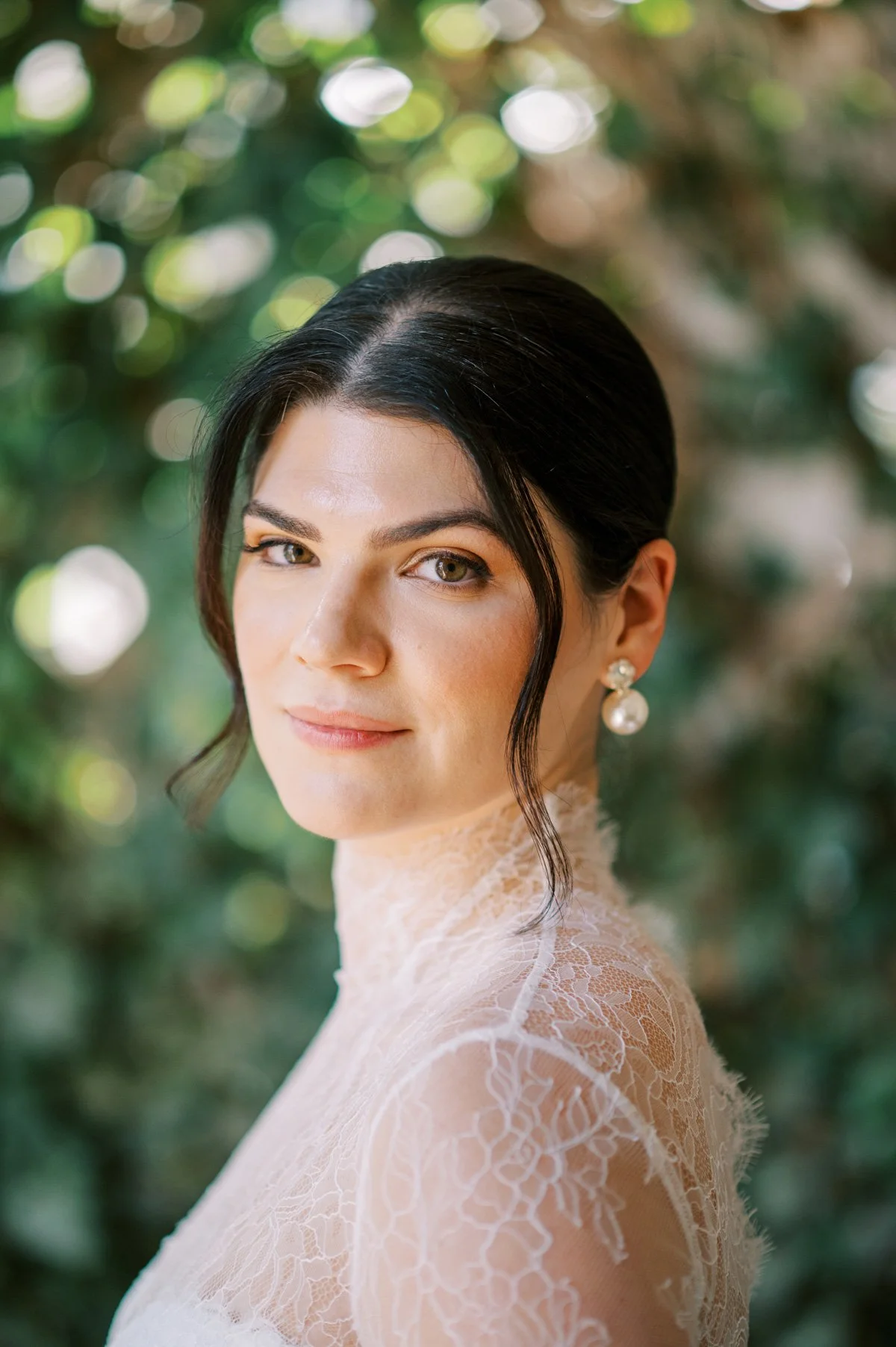 A woman with dark hair styled in a vintage updo, wearing pearl earrings and a lace top, standing outdoors with a bokeh background of green foliage.