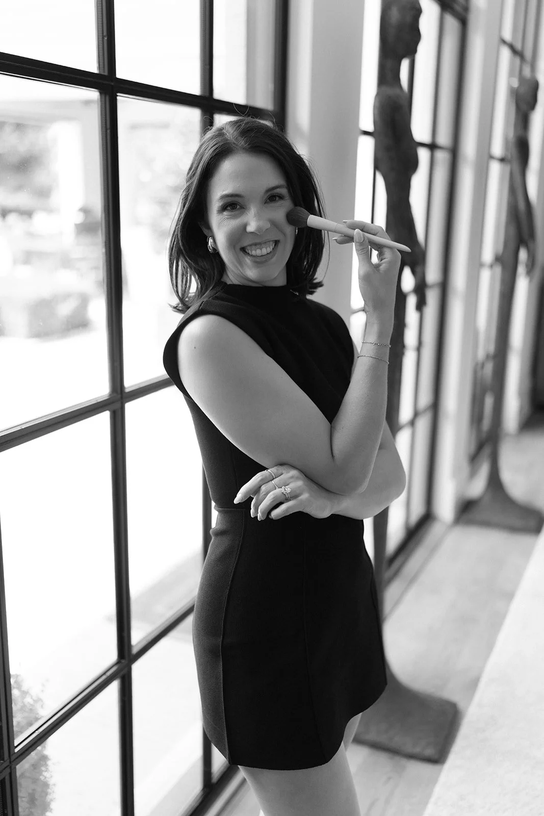 Black and white photo of Carly Marie smiling confidently while holding makeup brush by window