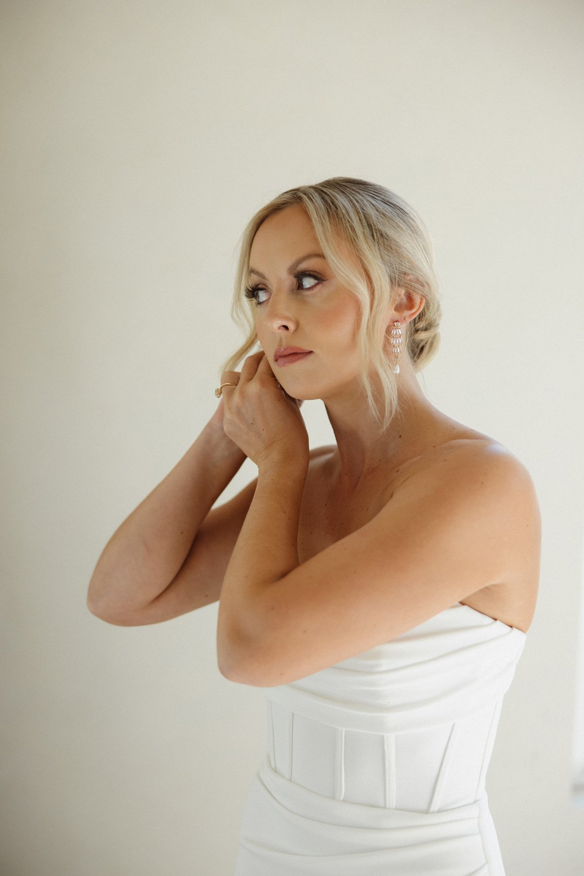 A woman with blonde hair styled in loose waves, wearing a strapless white dress and dangling earrings, looks thoughtfully to her left with one hand near her face against a plain, light-colored background.