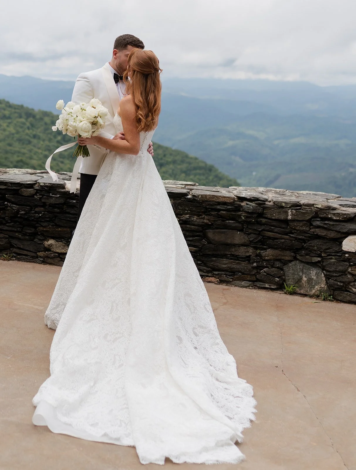 A bride in a white dress poses on the top of a mountain with her husband. Her hair is a half up half down bridal hairstyle