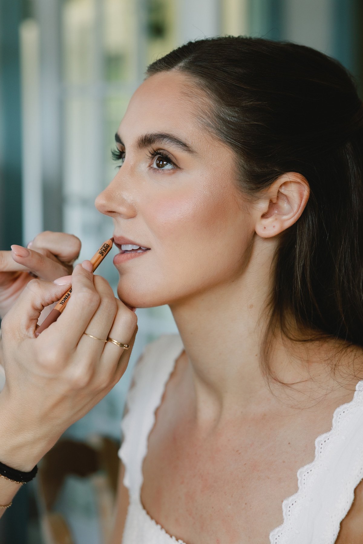 A woman with brown hair and light skin having her makeup done, with a makeup artist applying lip liner.