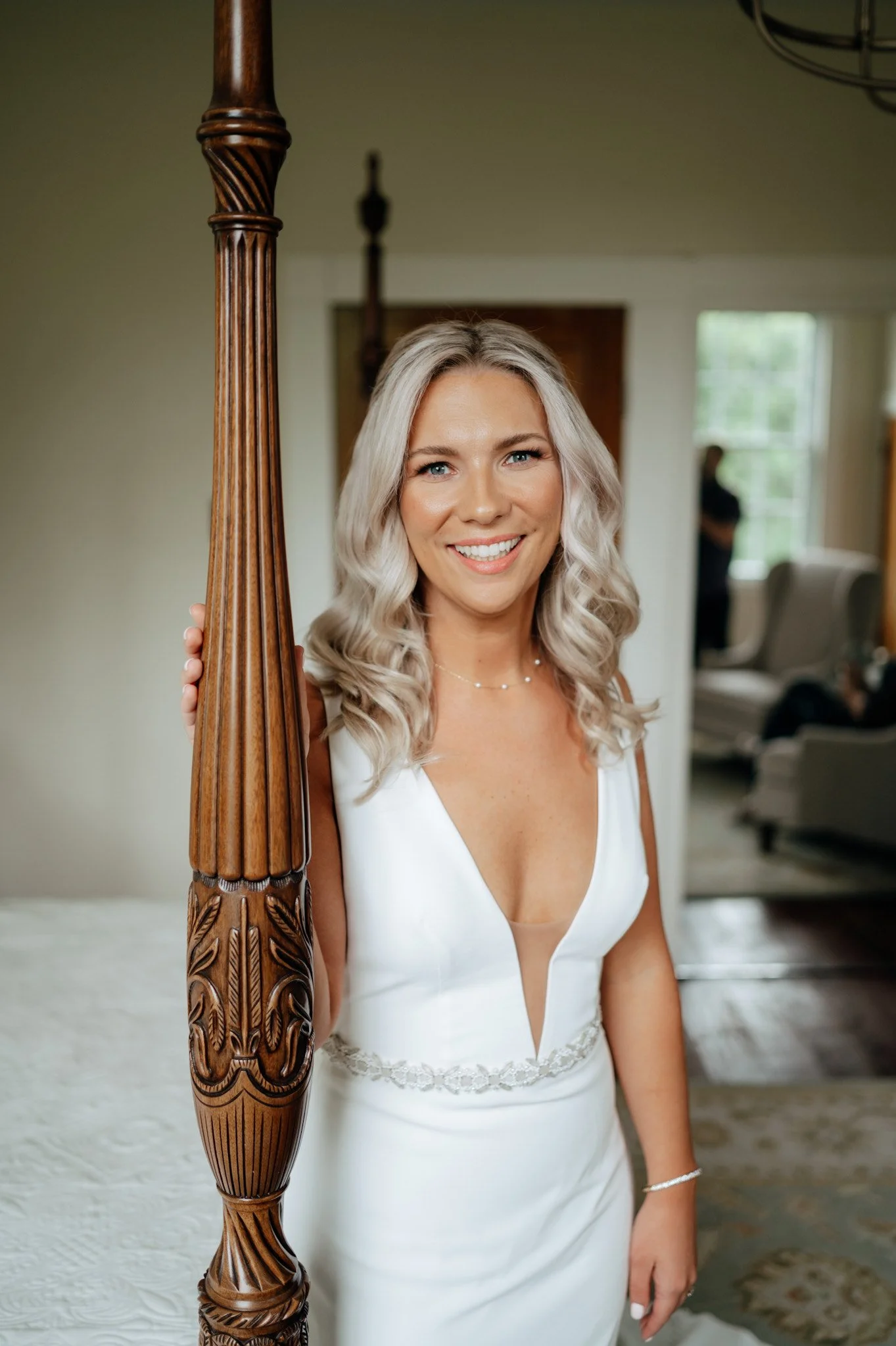 A woman in a white wedding dress smiling and holding onto a carved wooden bedpost inside a well-lit room.