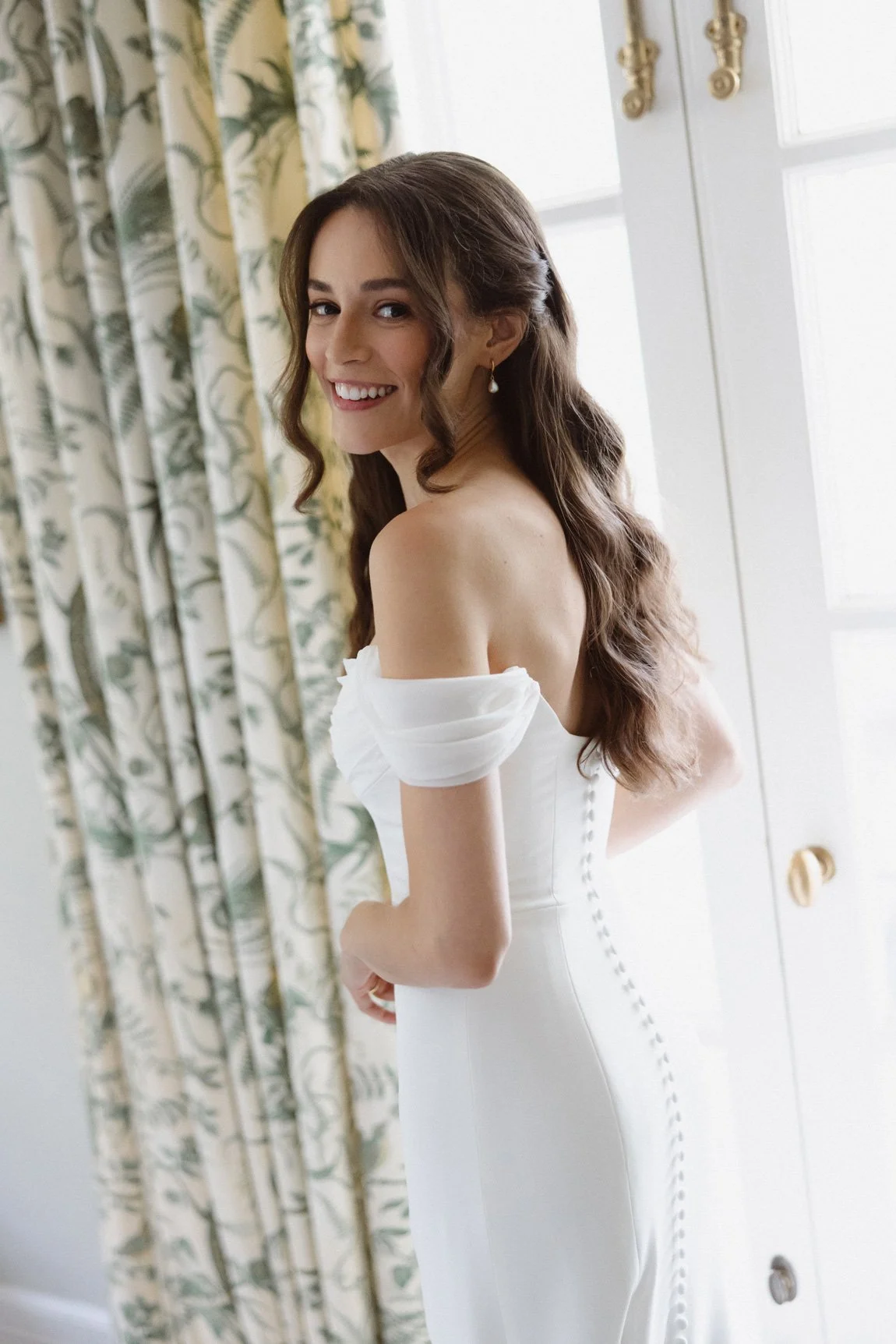 A woman in a white off-shoulder dress, smiling, standing near a window with patterned curtains.