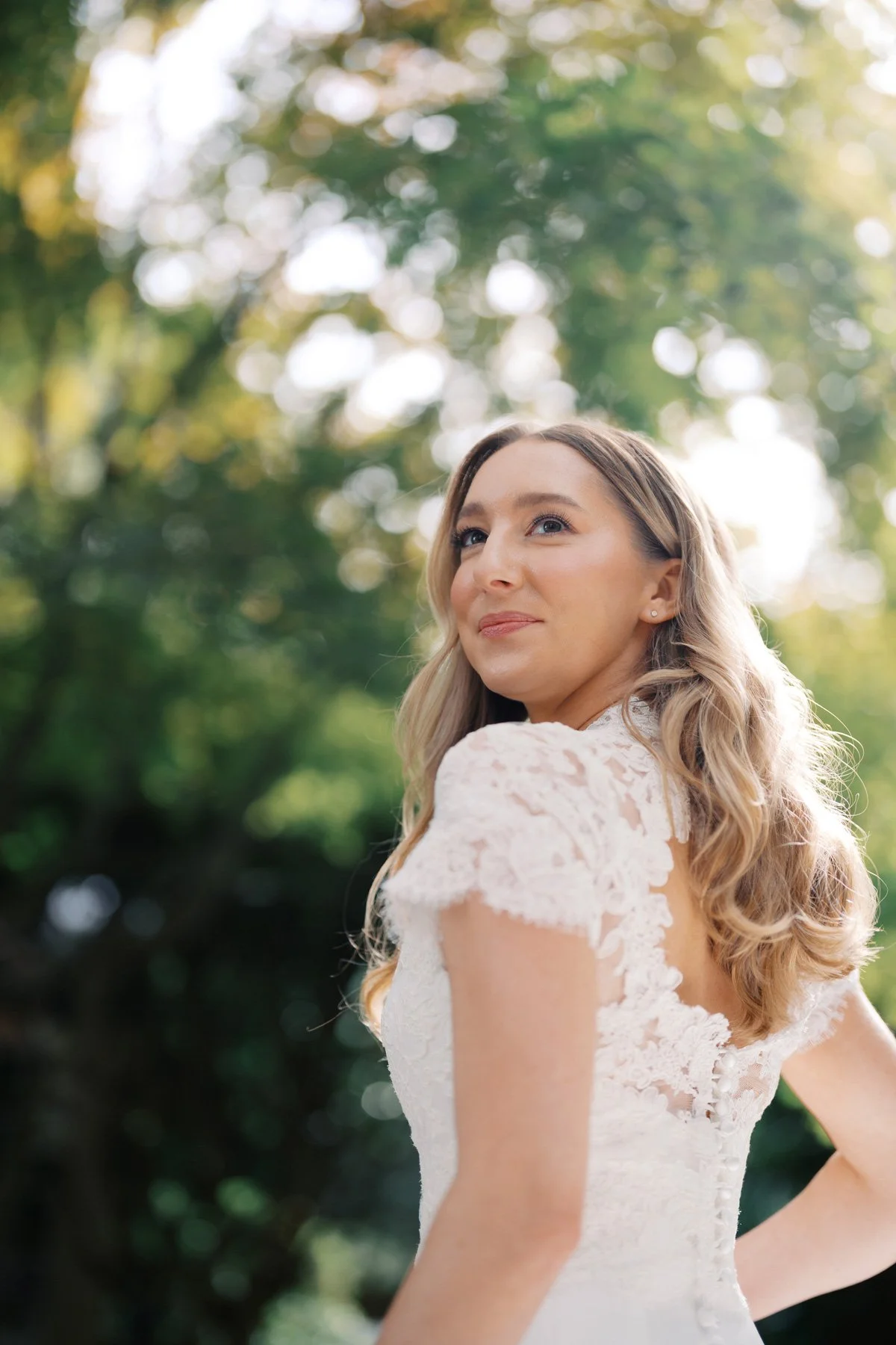 A woman with blonde wavy hair wearing a white lace dress stands outdoors among green trees, gazing to the side with a slight smile.