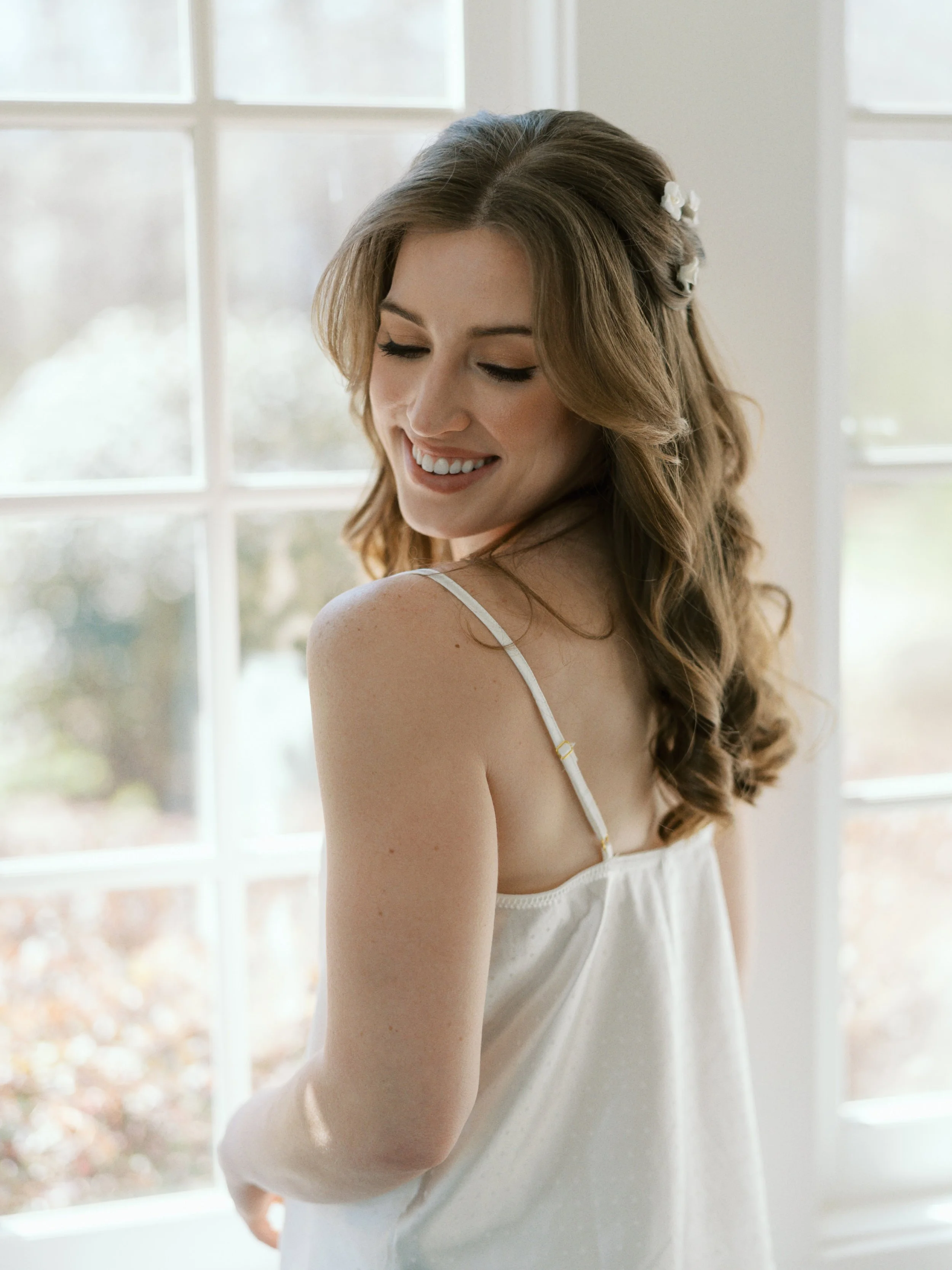 A woman with brown wavy hair looking over her shoulder in front of beautiful windows, waiting to get married. Bridal hair and makeup by professional makeup artist, Carly Amico of Carly Marie Artistry, a makeup artist in Charlotte NC.