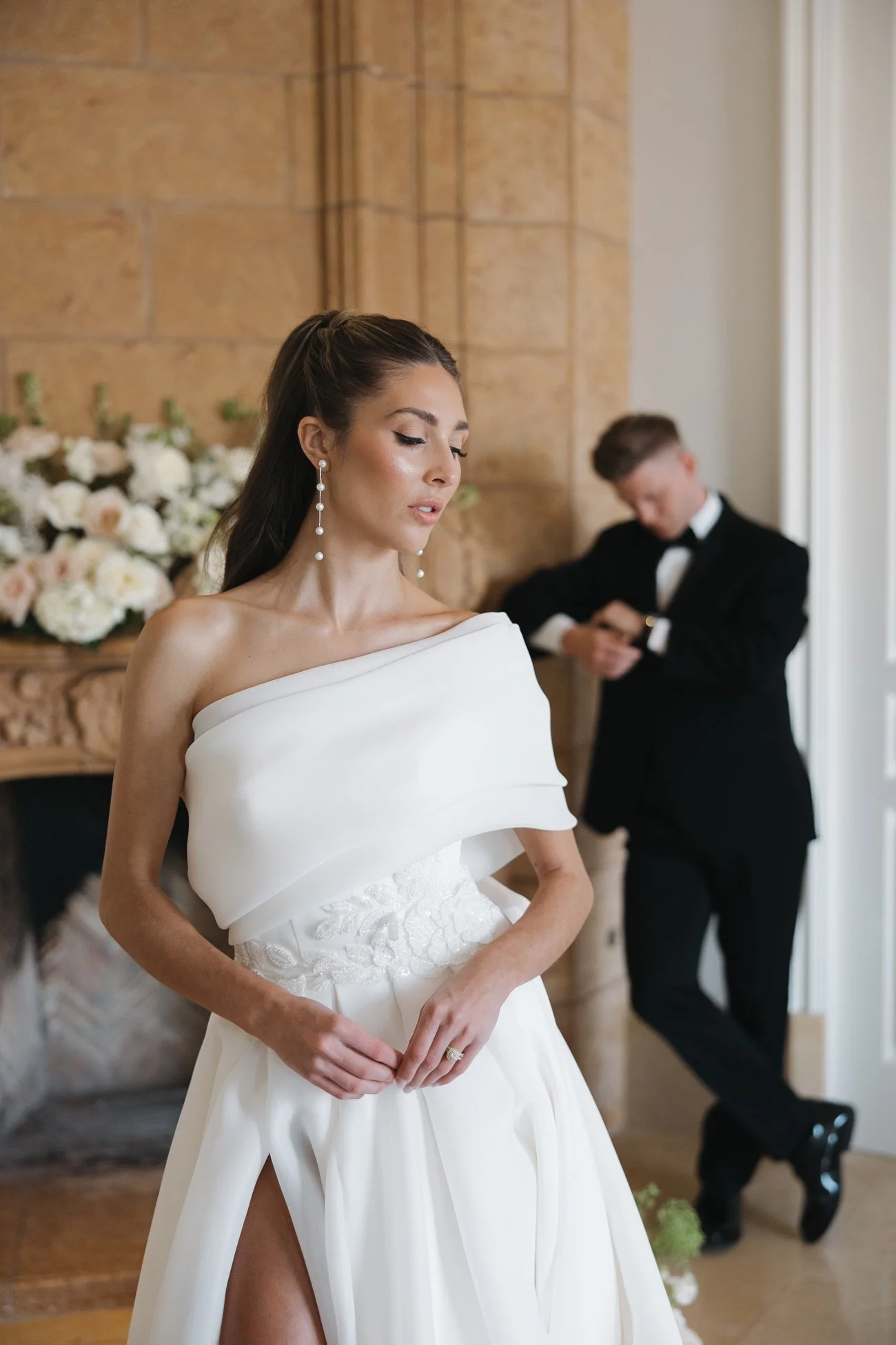 A bride in a white wedding gown with off-the-shoulder design and slit, standing with her hands clasped in front of her, looking down. Behind her, a groom in a black tuxedo with a bow tie is checking his watch near a floral arrangement by a fireplace.