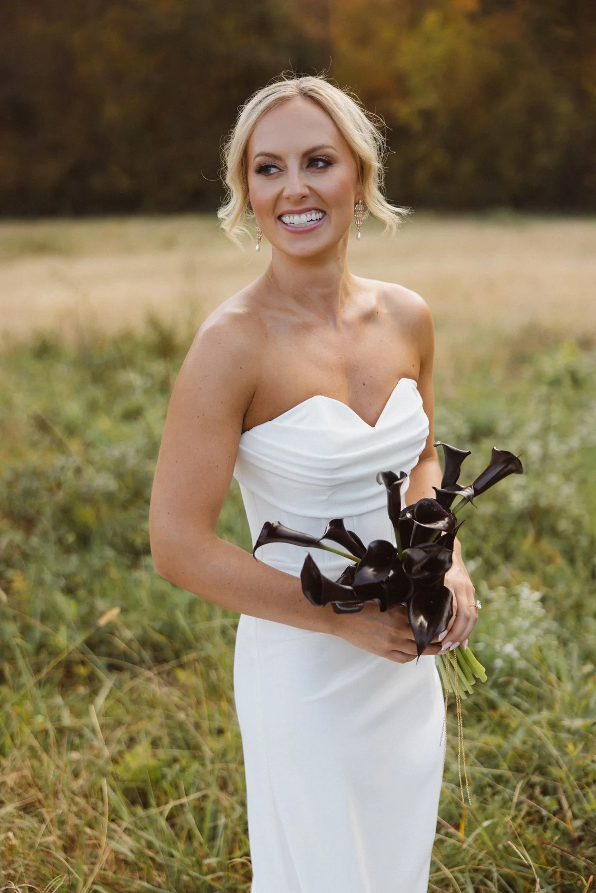 A woman in a white strapless dress holding a bouquet of dark purple calla lilies in an outdoor field during fall.