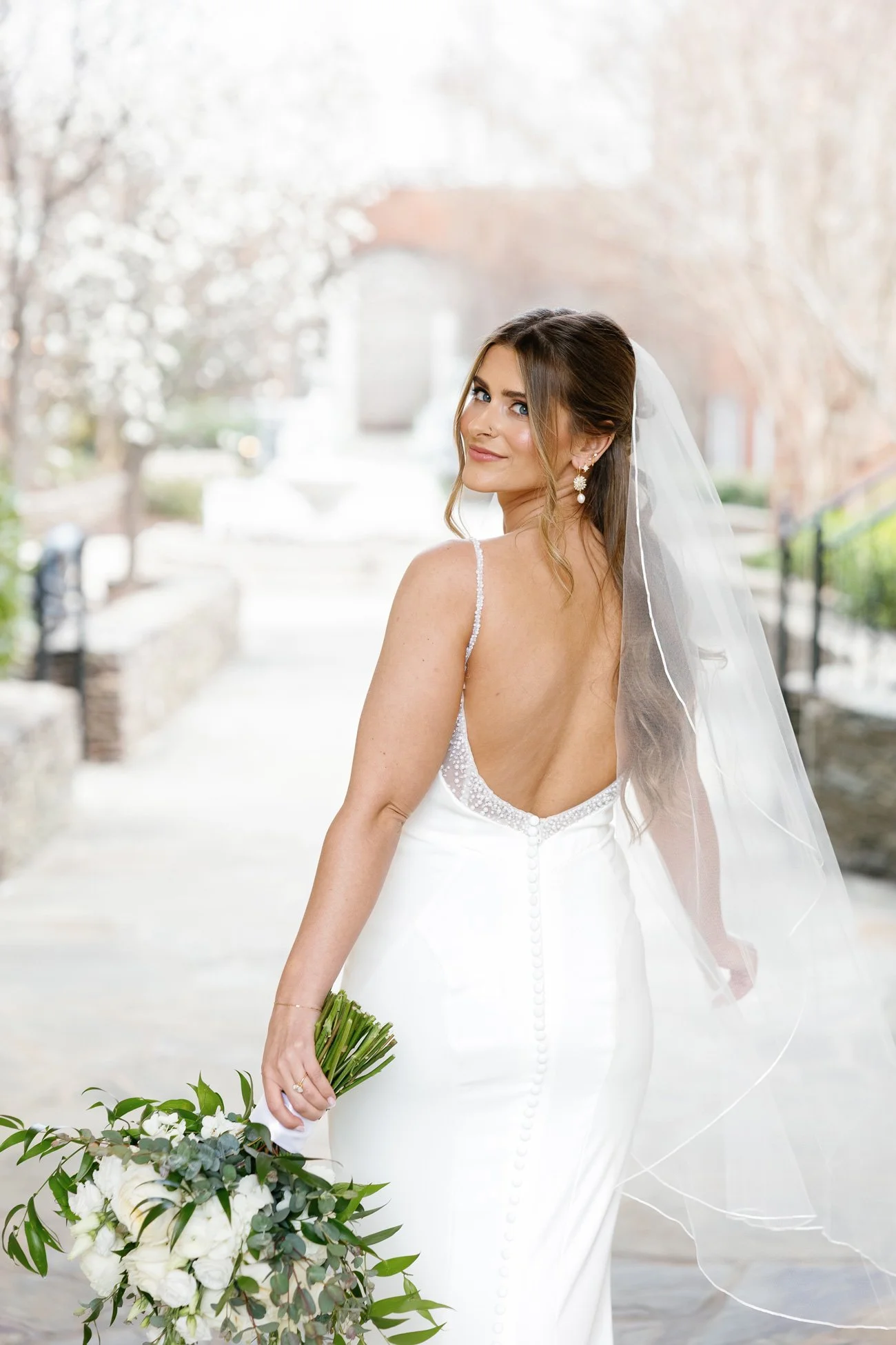 Bride walking away in fitted lace gown with low back and long veil, glowing with romantic bridal glam