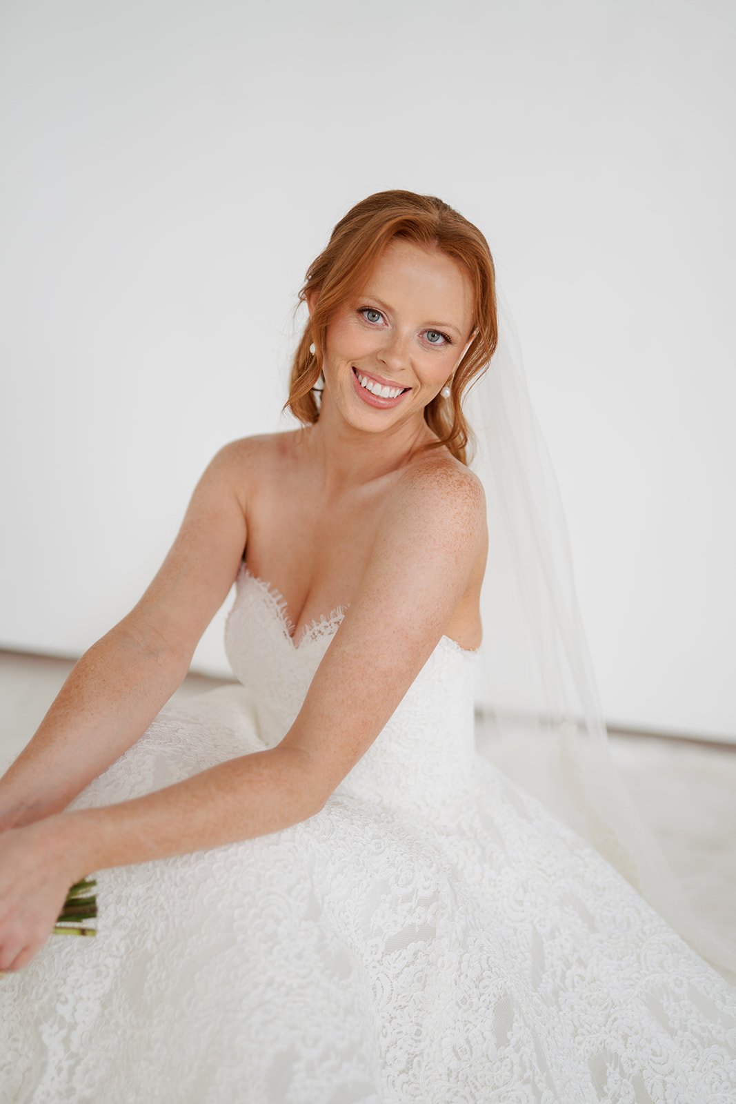 A woman in a white dress is a bride. Her hair is a half up half down bridal hairstyle. Her makeup is a soft glam bridal makeup look. She is holding flowers