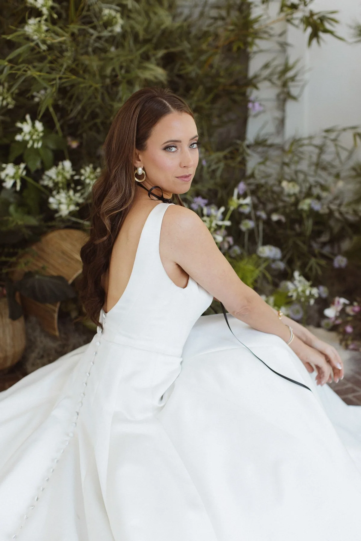A woman in a white wedding dress sitting against a backdrop of green plants and flowers, looking over her shoulder.