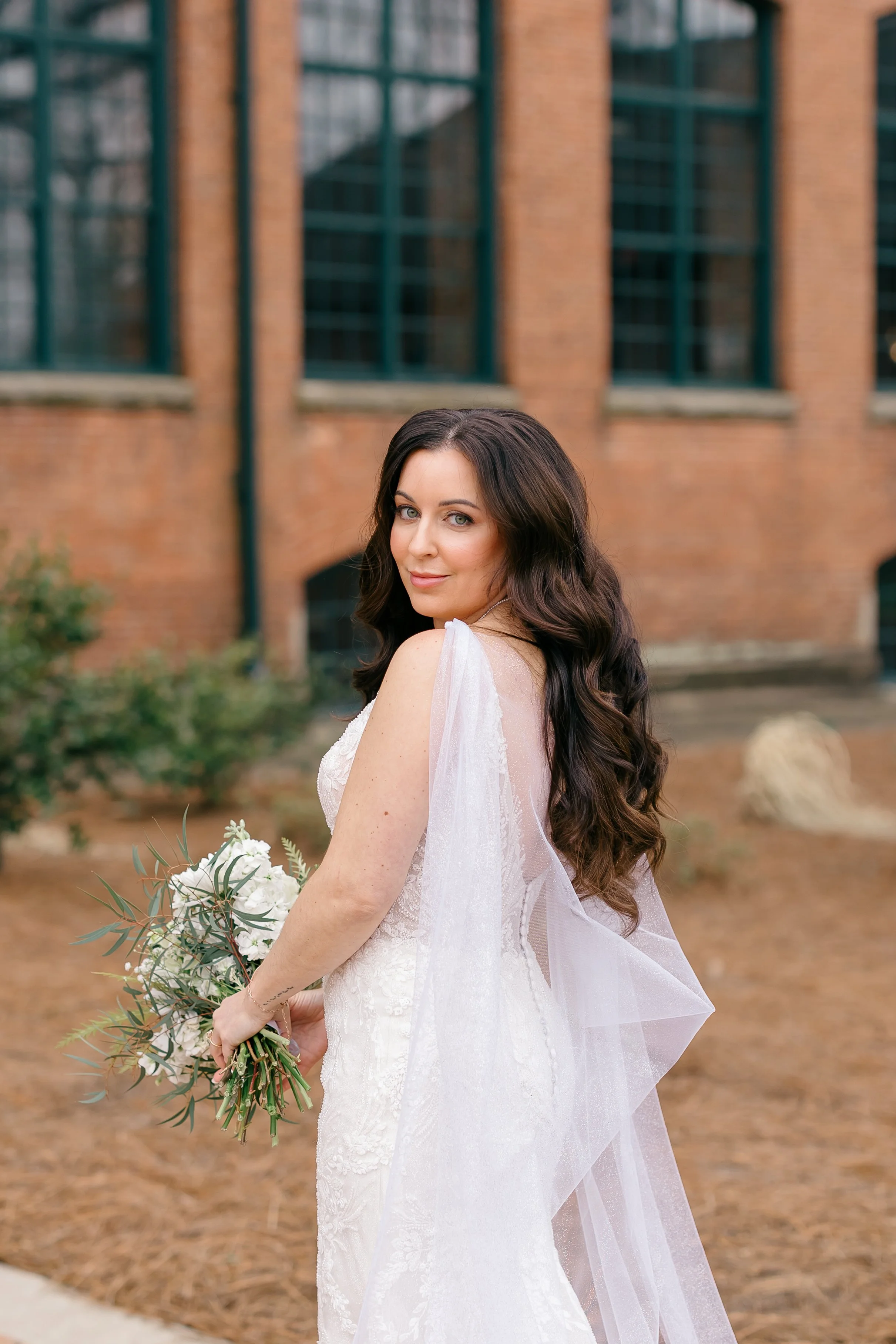 Bride standing confidently in front of brick venue with soft waves and bridal glam makeup