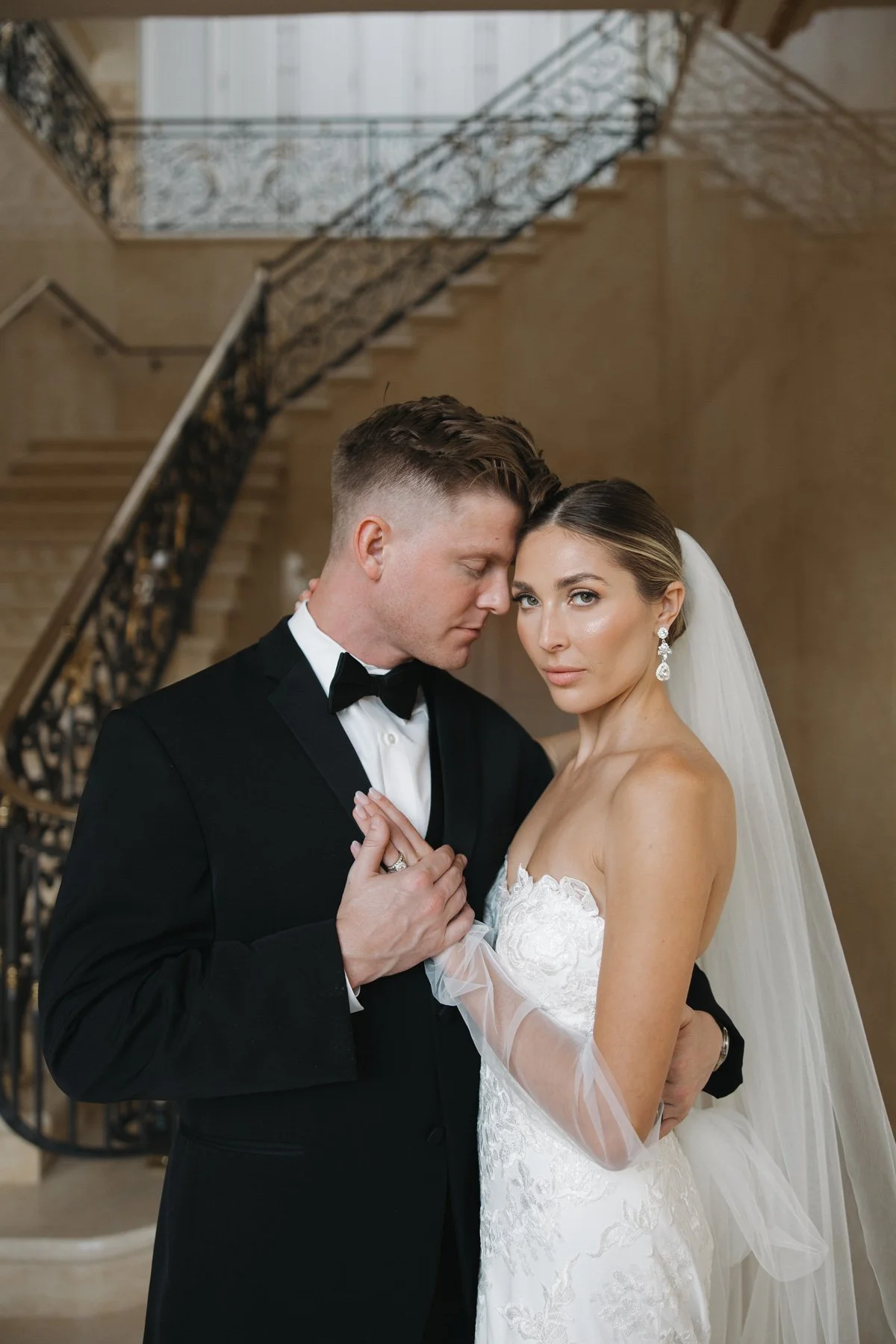 A bride and groom in wedding attire, embracing in front of a staircase inside a building.