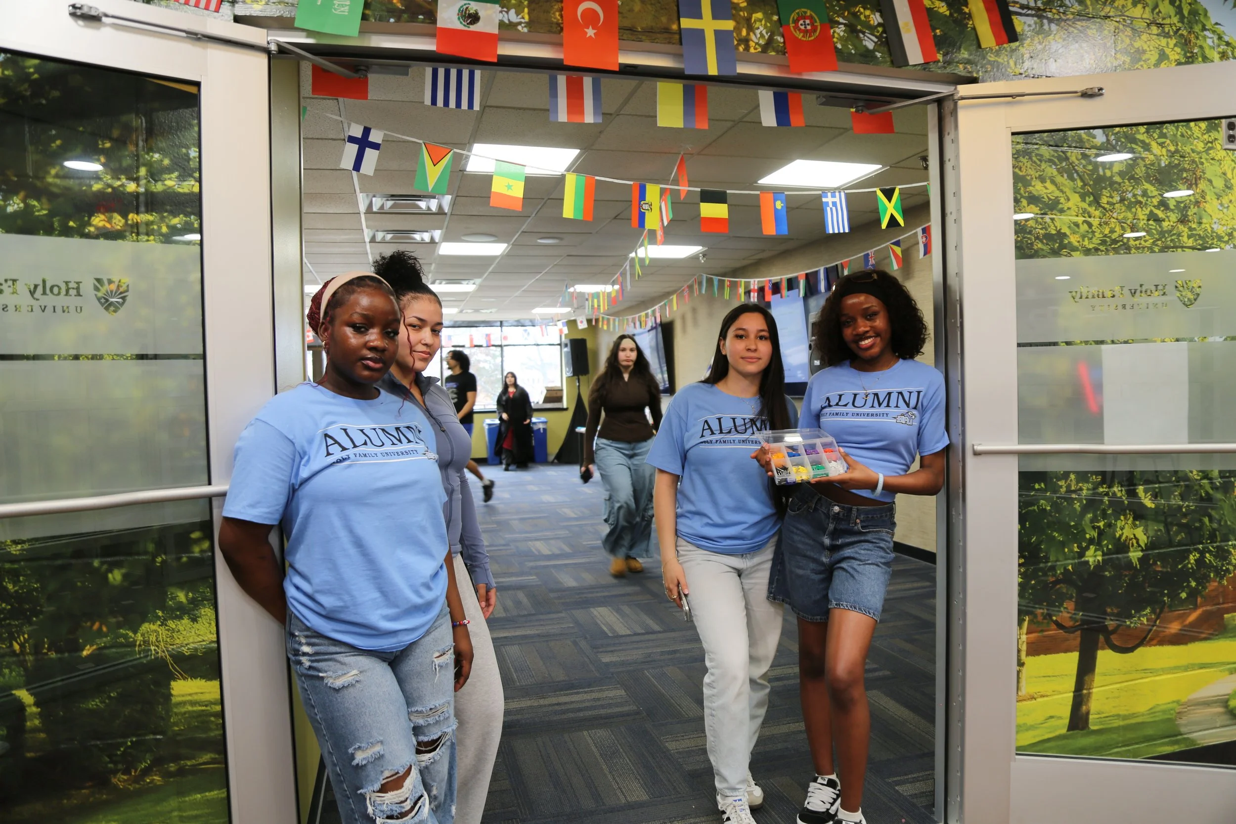 Four young women standing in a hallway decorated with international flags, three wearing light blue 'Alumni' t-shirts, two of them smiling, one of them holding a small plastic container of candy, in a university setting.