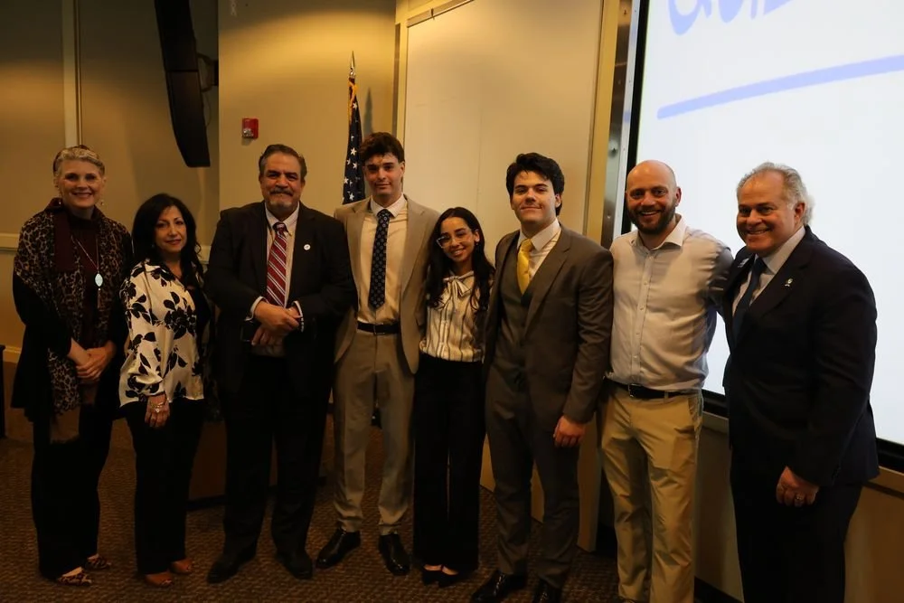 Group of nine people standing together indoors, posing for a photo at a formal event with a projector screen and a flag in the background.