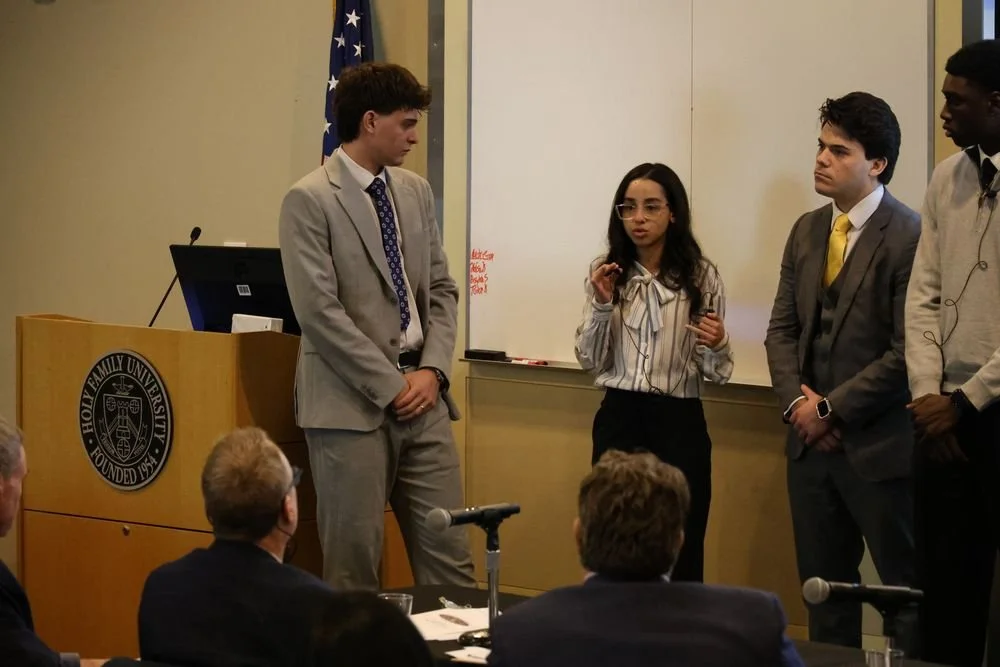 A group of four students is standing at the front of a classroom, giving a presentation to an audience. One girl is speaking, holding a microphone, while the three boys listen. There is a whiteboard behind them and an American flag in the room. The students are dressed professionally.