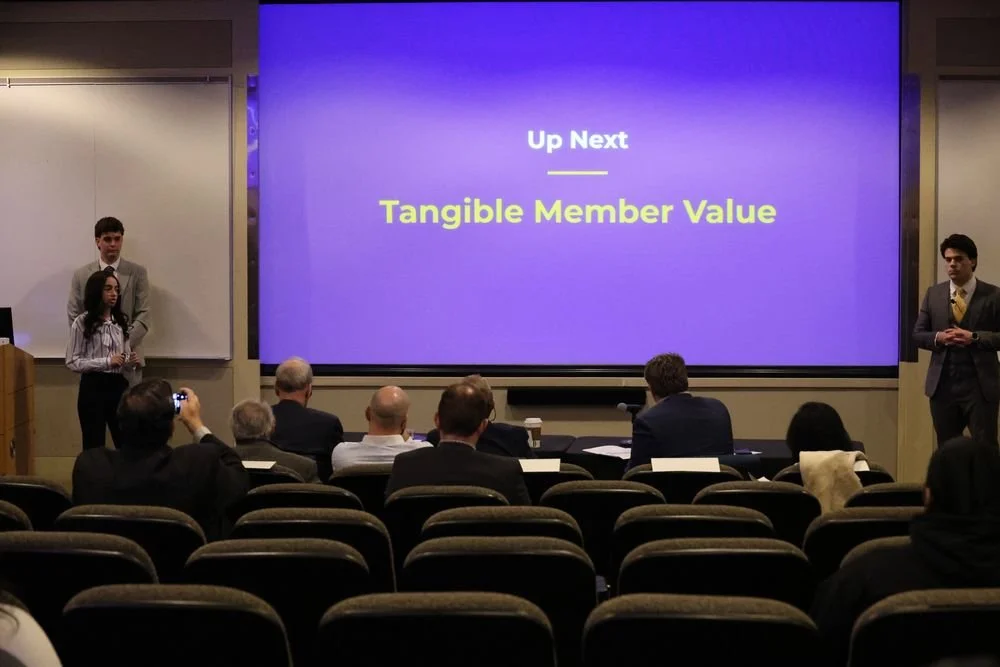 Presentation in a conference room with three presenters and an audience, showing a slide titled 'Tangible Member Value' with ‘Up Next’ at the top.
