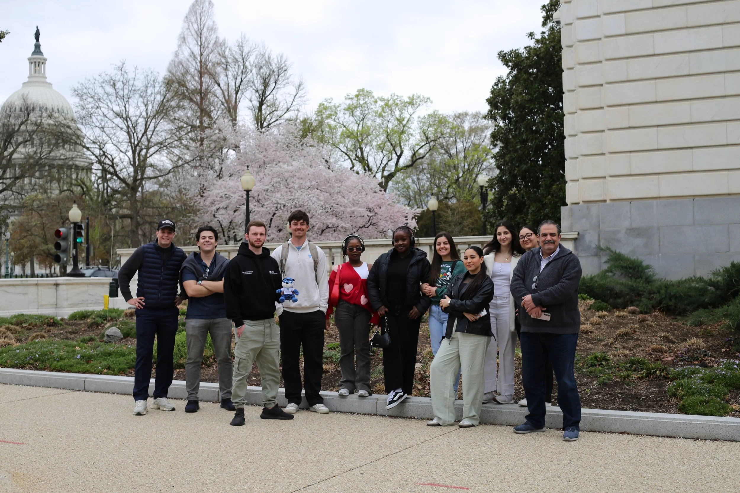 Group of ten diverse people standing outdoors in front of a flowering cherry blossom tree, Capitol building, and a stone building, with some holding a plush toy, during daytime with overcast sky.
