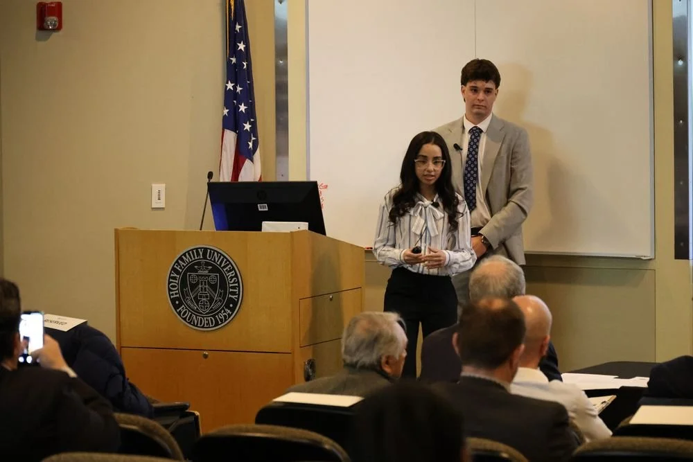 Two young individuals stand at the front of a lecture hall, giving a presentation. The woman is holding a microphone and wearing glasses and a striped blouse. The man is wearing a light gray suit and has a serious expression. An American flag is behind them, along with a whiteboard, and they are presenting to an audience of seated adults.