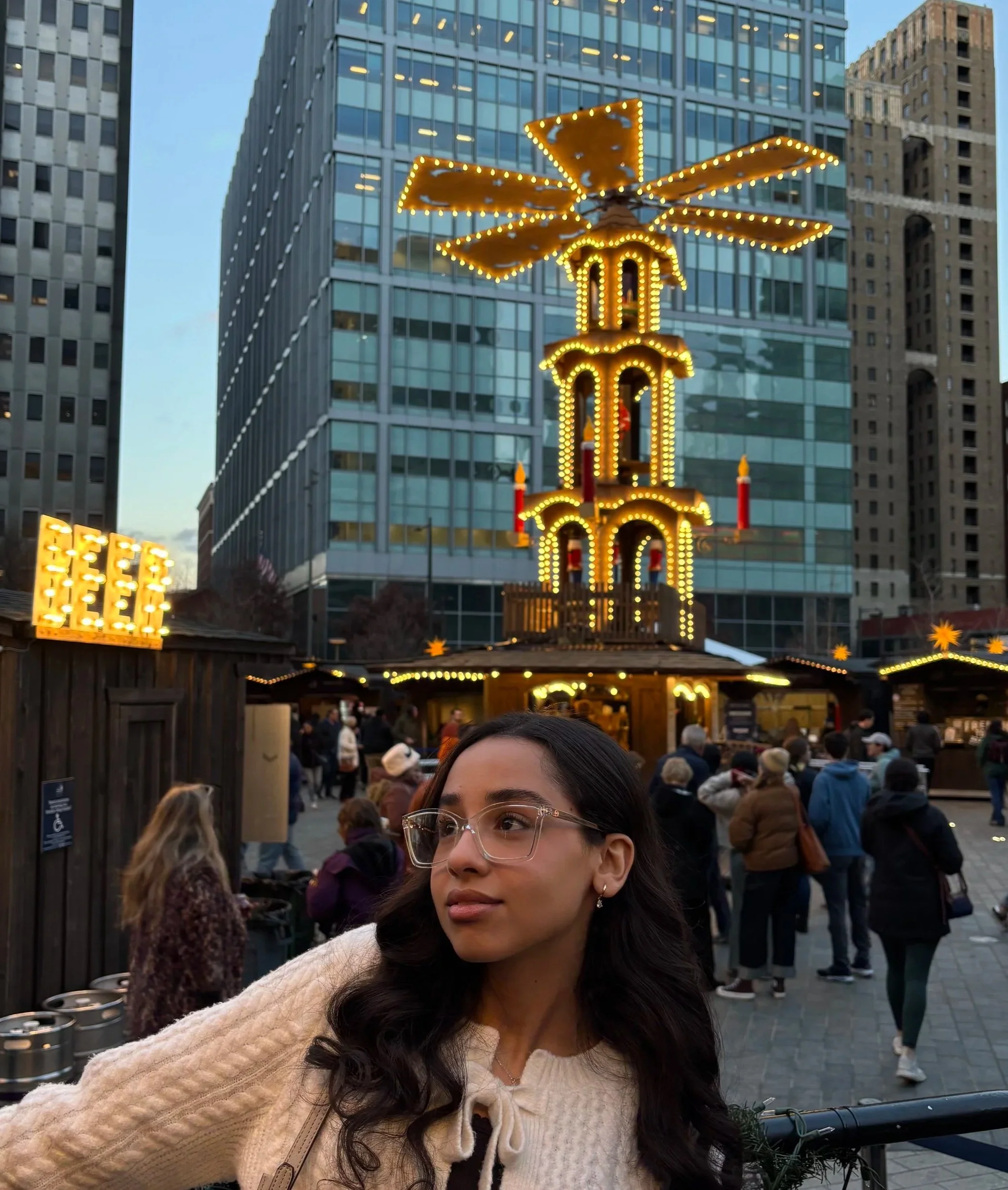 A woman with glasses and long dark hair stands in front of a festive outdoor market with a large illuminated gingerbread multi-tiered star and city skyscrapers in the background.