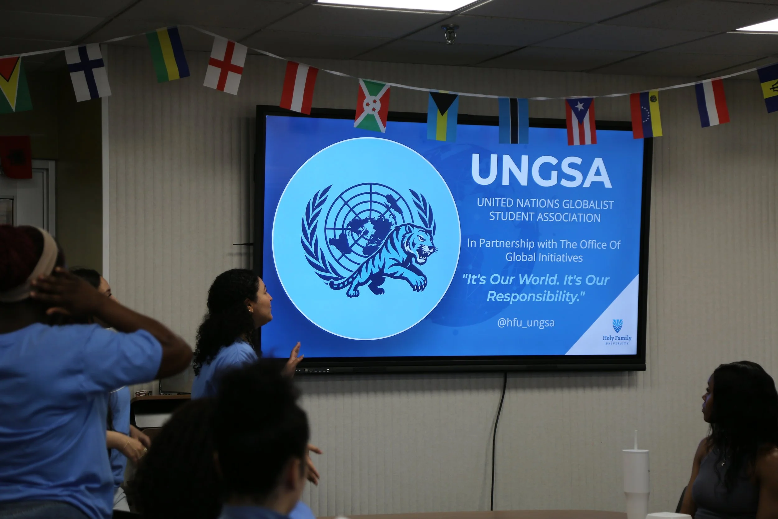 Students attending a meeting or presentation for the UNGSA (United Nations Globalist Student Association) at Holy Family University, with a large screen displaying the organization's logo, mission statement, and social media handle, decorated with international flags.